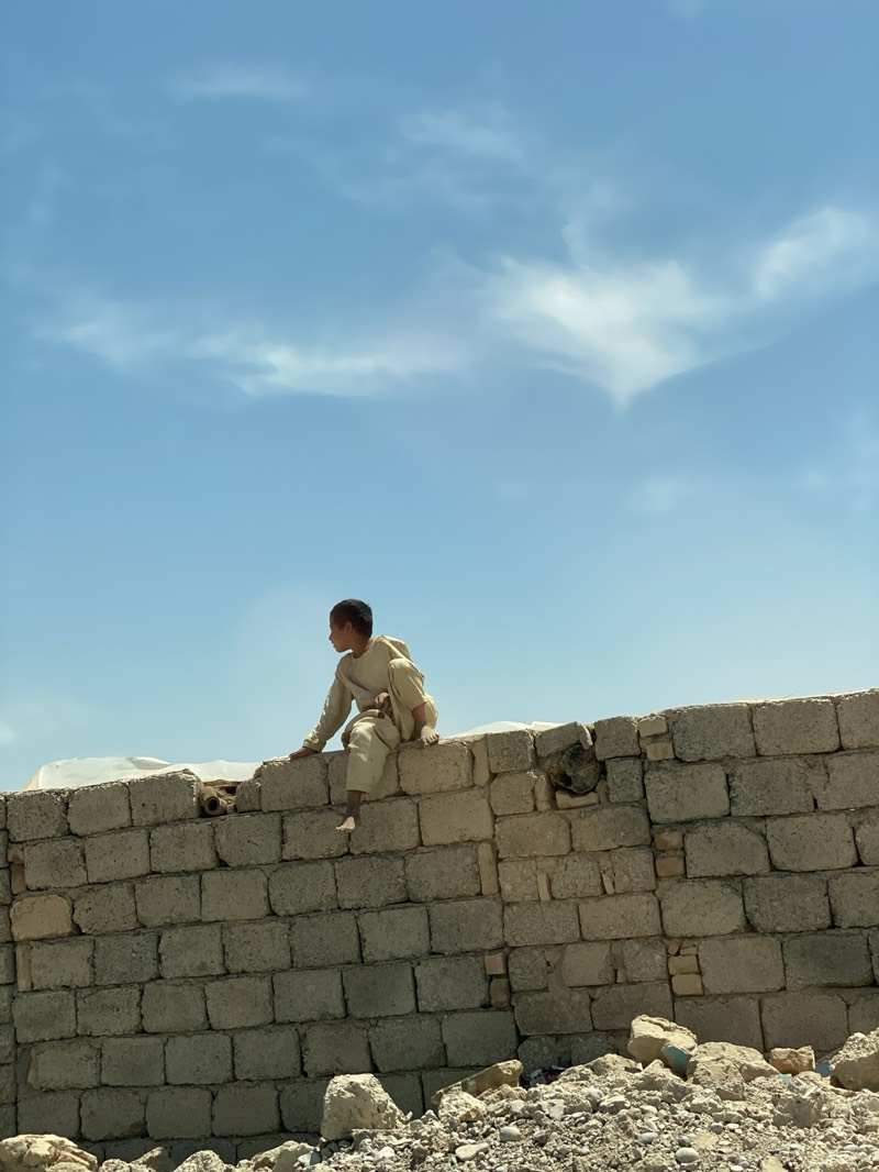 A young boy in a light-colored outfit sits on top of a low brick wall in Deh-E Bagh, Afghanistan. The wall is made of gray bricks, and there is debris and rubble at its base. The sky is light blue with some clouds. The boy is looking to the right of the frame.