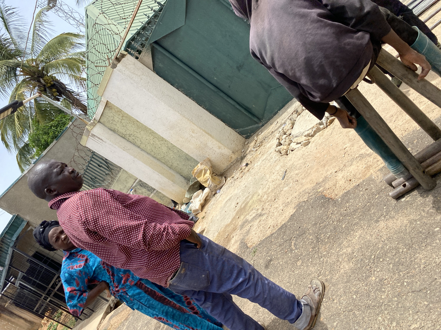 This outdoor image, captured from a tilted perspective in Jos, Nigeria, shows several people and urban architectural elements under bright daylight.
In the foreground and center, a bald, dark-skinned man in a maroon shirt with white polka dots and blue jeans stands, looking upwards and slightly to his right. His left hand is in his pocket, and he wears light-colored, possibly worn, shoes. Partially visible behind him is a woman with a dark headscarf, wearing a vibrant blue patterned dress, also standing.
To the far right, another individual is seen from behind, wearing a dark-colored top (possibly a hoodie) and sitting on a makeshift wooden structure, which appears to be a crude ladder or scaffolding. Their legs are clad in green boots or pants, and their hands are resting on the wooden rungs.
The background features a light-colored concrete wall topped with barbed wire, suggesting a security measure. Behind the wall, a tall, corrugated green metal gate is visible. Above the gate and wall, the fronds and coconuts of a palm tree are prominent against a clear, bright sky. The ground beneath the subjects is a mix of concrete and dirt, uneven, with scattered debris including small rocks and a crumpled bag or paper. No discernible text is visible in the image. The scene suggests a casual gathering or a moment in a busy urban or semi-urban setting.