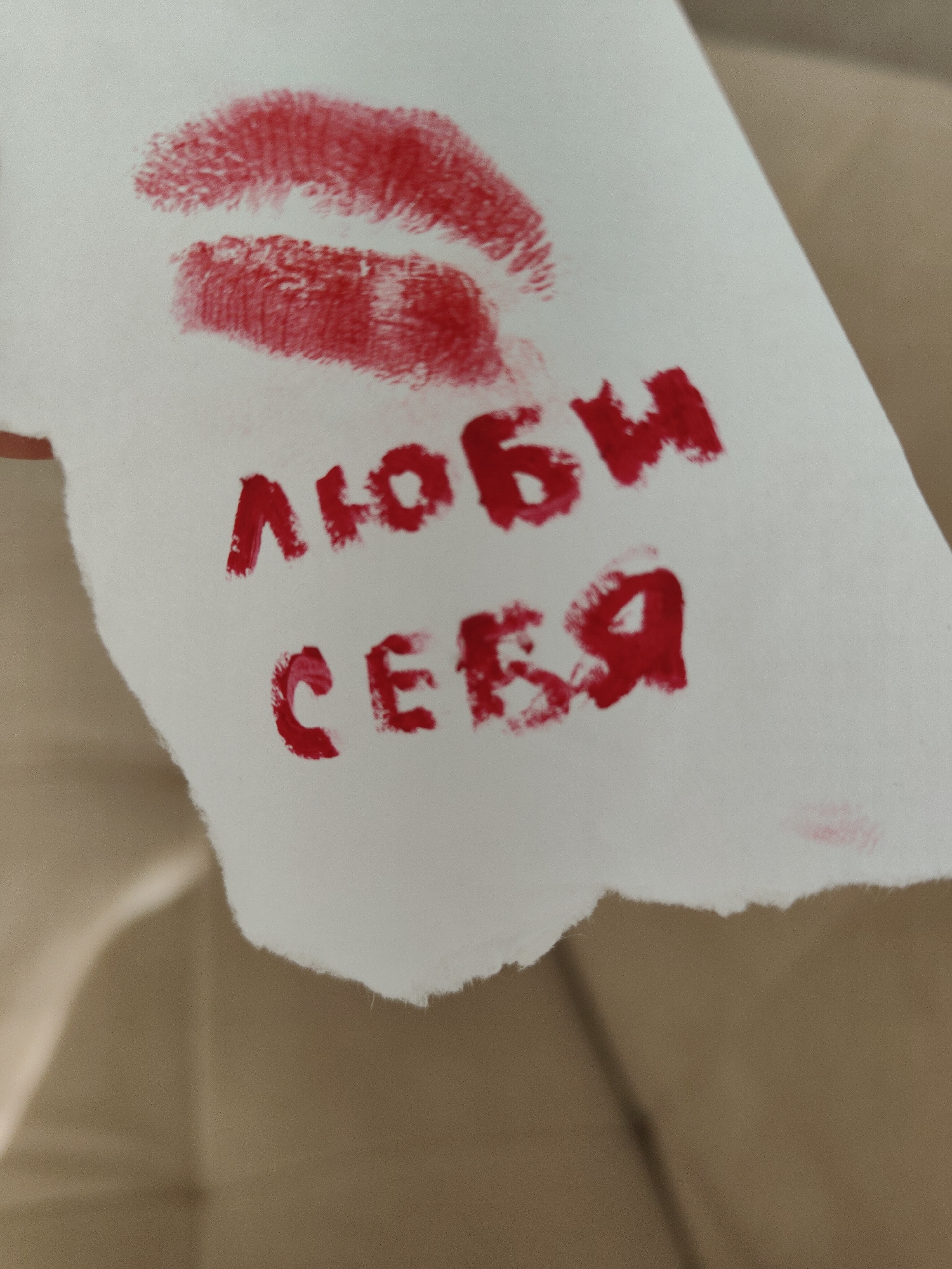 A white piece of tissue or cloth is held against a blurred tan background, likely fabric from a piece of furniture. There is a red lipstick print at the top of the tissue. Below the print, the words "ЛЮБИ СЕБЯ" are written in red marker or paint. The location and other specific details are not discernible from the image.