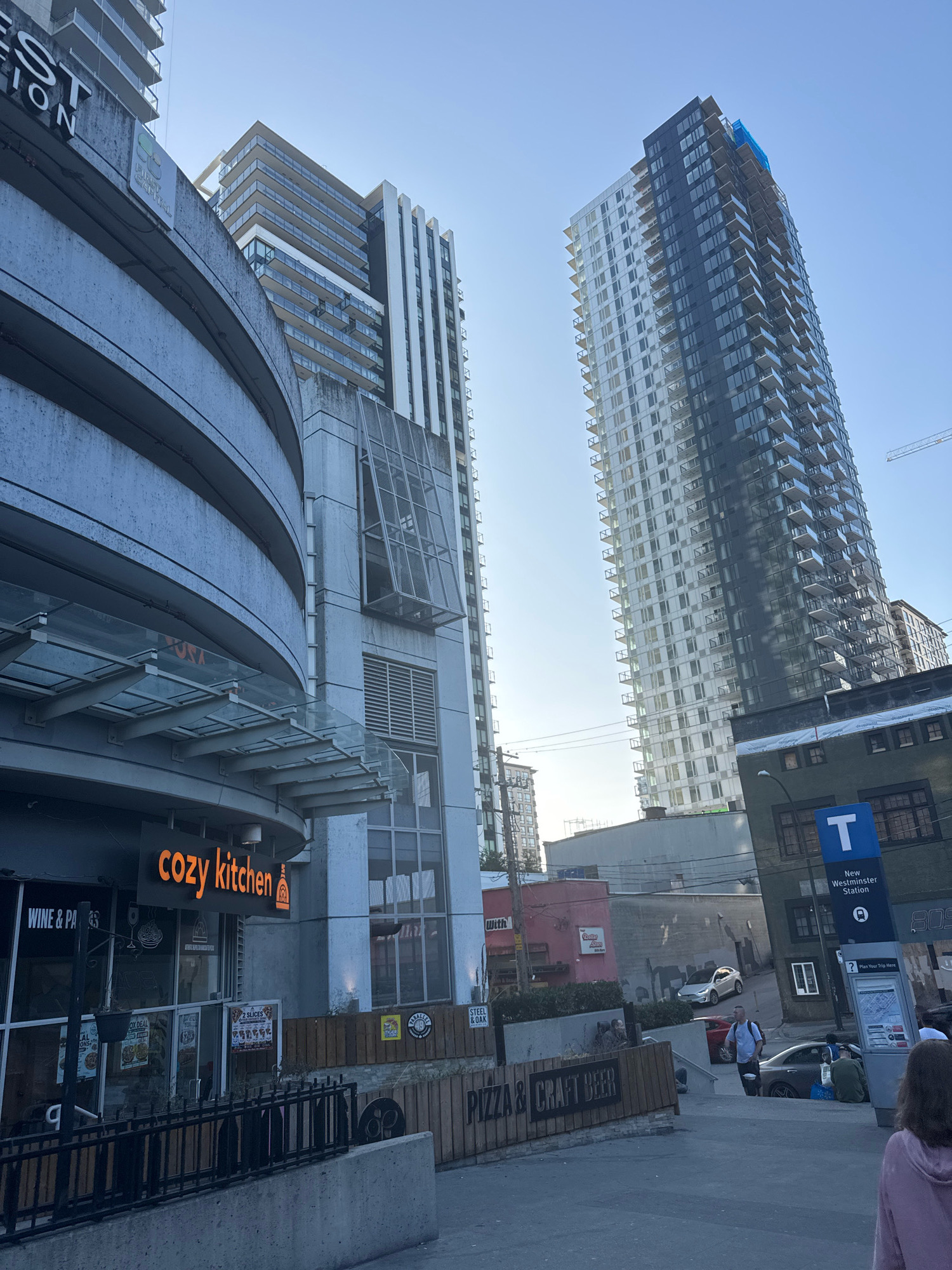 The image shows an urban street scene in New Westminster, Canada, dominated by modern high-rise buildings and commercial establishments. In the foreground, a curved concrete structure with a glass awning leads to a storefront with a neon sign reading "cozy kitchen." Next to it, a sign indicates "WINE & PA". A black metal fence runs along the edge of a raised walkway. Further into the scene, two tall residential buildings stand out: one is primarily white with glass balconies, and the other is a darker, sleeker design with a dark facade and glass balconies. A blue "T" sign with "New Westminster Station" printed below indicates a transit stop. Several cars are visible on the street, and a few people are scattered throughout the scene, some walking and others sitting. The environment appears to be a commercial and residential area with a clear sky overhead.