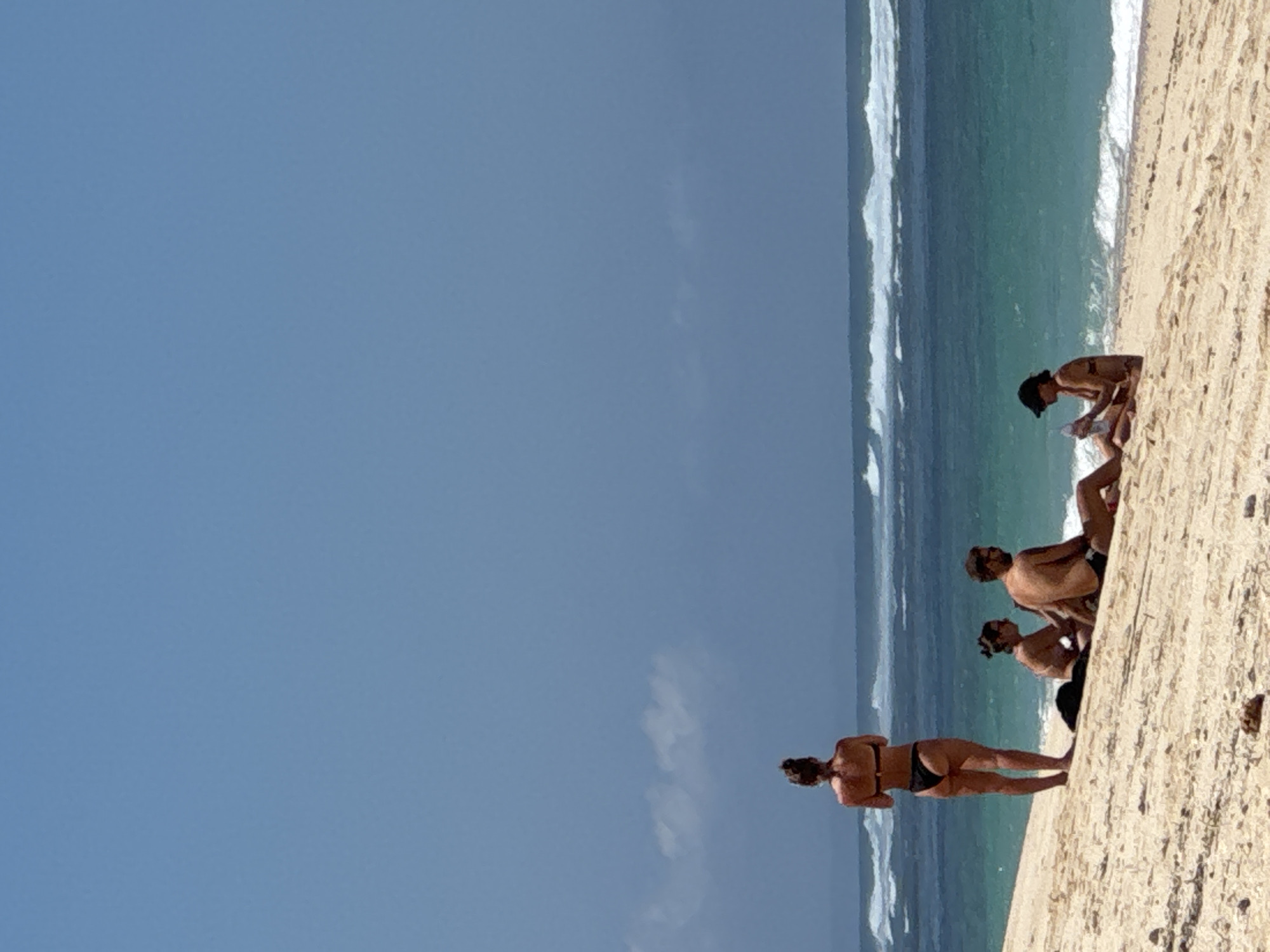 On a sandy beach in Kuta Selatan, Indonesia, four individuals are present near the water's edge. A woman stands facing away from the camera, looking out at the ocean. To her left, a man and a woman are seated on the sand. Further to the right, another person is seated, holding what appears to be a bottle. The ocean is visible with waves breaking, and the sky is a clear blue with some scattered clouds. The sun is shining, creating a bright atmosphere.