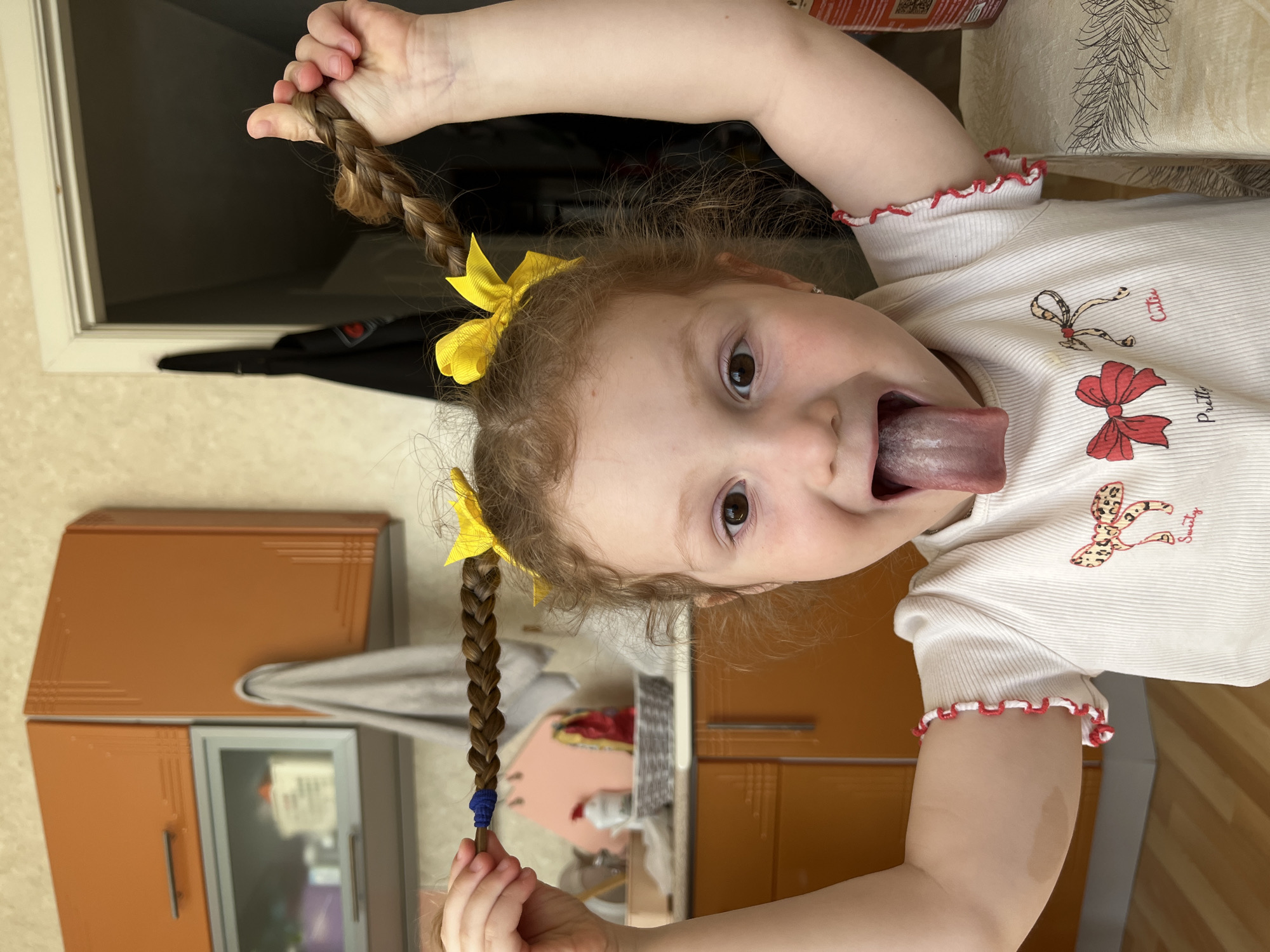 The image depicts a young child, likely female, with auburn, curly hair styled into two braided pigtails, each adorned with a yellow bow. The child is positioned centrally in what appears to be a home kitchen or dining area in the city of Chaikovsky, Russia. She is actively posing with her arms raised, holding one braided pigtail in each hand. The pigtail on the viewer's left is secured with a blue elastic band, while the one on the right is held directly by her hand. She is making a playful expression, sticking her tongue out. Her brown eyes are open and directed towards the viewer. She wears a white, short-sleeved top with ruffled red trim on the sleeves and embroidered bows on the chest, accompanied by the text "Pretty" and "Cutie." A red and white patterned bracelet is visible on her right forearm.
The background reveals a domestic environment. To the viewer's left, orange kitchen wall cabinets with silver handles are mounted above a lower cabinet featuring a frosted glass door. Below these, a countertop holds a pink cutting board, a white teapot with a red lid, a woven basket, and a red package. To the viewer's right, a light-colored table with a dark, patterned tablecloth is partially visible. Further right and behind, an open doorway leads to a darkened room, with a dark coat hanging on a hook beside the door frame. The walls are covered in light-colored, textured wallpaper. A wooden floor is visible in the lower-left background.