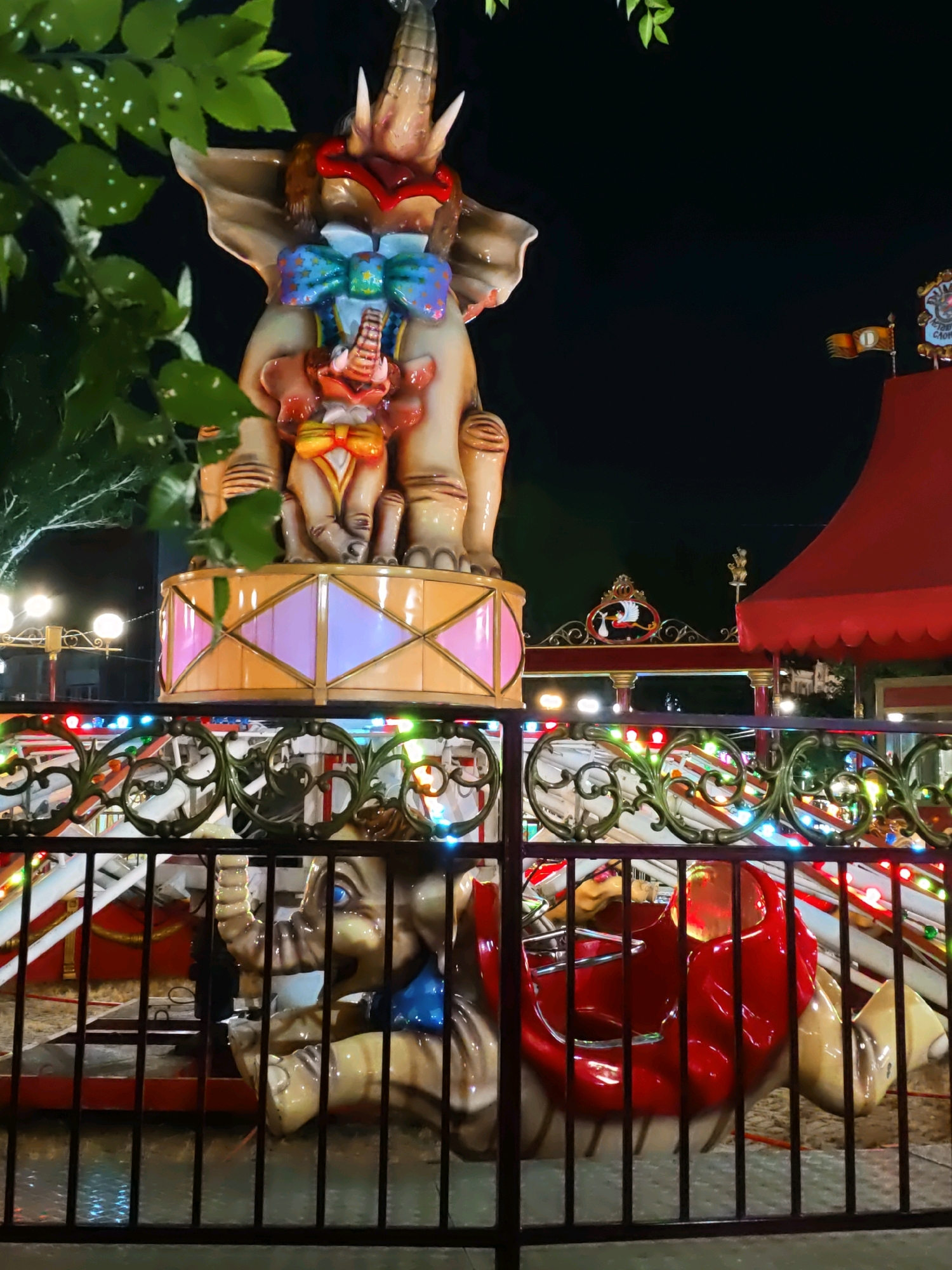 This is a nighttime outdoor photograph of an amusement park ride in Rostov-on-Don, Russia. The ride features decorative elephant figures, with a large, ornate elephant at the top of the frame, adorned with a colorful bow and a smaller elephant figure. Below, another elephant figure, serving as a seat for the ride, is visible behind a black metal fence. The ride is illuminated by colorful lights and is situated near a red tent-like structure. The background is dark, indicating it is nighttime.
