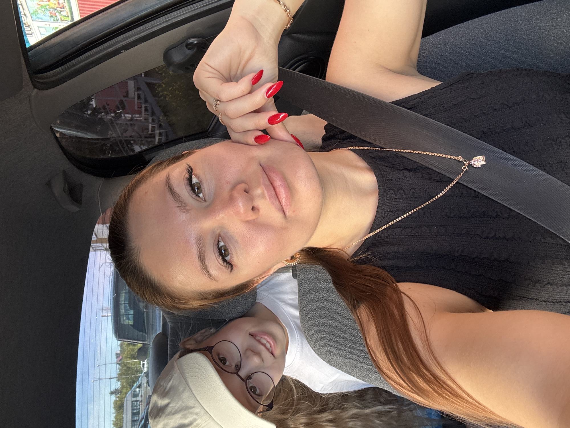 In the foreground of the image, a woman with long, auburn hair is seated in the driver's seat of a car, taking a selfie. She is wearing a black sleeveless top and a gold chain necklace with a small pendant. Her right hand, with bright red fingernails, is resting on her cheek. A child, wearing glasses and a white t-shirt, is visible in the passenger seat behind her, wearing a light-colored baseball cap. The child is looking towards the camera and smiling. The interior of the car is visible, including the gray seats and a black seatbelt across the woman's chest. The background, seen through the car's windows, shows a blurred outdoor scene with trees, buildings, and other vehicles, suggesting the car is in a city environment. The lighting suggests it is daytime, possibly late afternoon due to the warm tones. The overall scene depicts two individuals inside a vehicle, likely on a journey.
