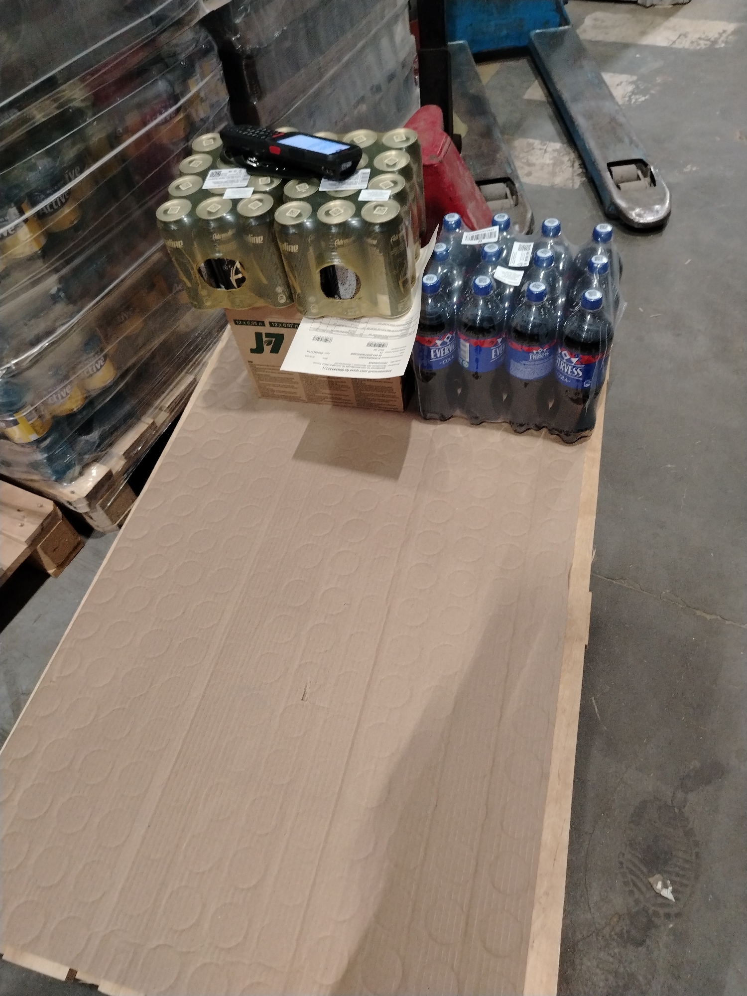 An overhead view captures a section of what appears to be a warehouse or storage facility in Zavolzhye, Russia. The scene is dominated by products placed on a wooden pallet that is partially covered with a large sheet of corrugated cardboard.
On the cardboard, two main types of beverages are visible. To the left, a multi-pack of green and gold cans, shrink-wrapped in clear plastic, is present. The cans are branded "Adrenaline" and "Adrenaline Rush." A black and red handheld scanner with a lit screen is positioned on top of these cans, suggesting an inventory or scanning operation. A label on the can packaging indicates "12 x 0.55 л" (12 x 0.55 liters). Below the cans, a brown cardboard box with "J7" prominently displayed is partly visible, also featuring text "12 x 0.55 л 12 x 687 л."
To the right of the cans, another multi-pack of dark-colored bottled beverages, also shrink-wrapped, is visible. Their labels clearly show the brand "EVERVESS - COLA."
In the background, the metal forks and wheels of a pallet jack or similar industrial lifting equipment are visible against a concrete floor. To the far left, a large, tall stack of items, possibly more beverages, is wrapped in clear plastic film. Through this wrapping, labels like "Active" and "Adrenaline" are discernible. A white paper, likely an inventory or shipping document, with text "ный лист груза ва 000394375/1" is visible on the cardboard box beneath the cans.
The arrangement of goods, the scanner, and the pallet jack suggest an ongoing activity related to warehousing, stock management, or order fulfillment.