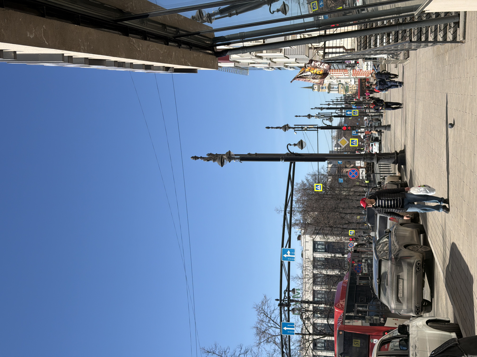 The image depicts a street scene in Perm, Russia, under a clear blue sky. The camera is oriented vertically. Along the right side of the frame is a sidewalk paved with light-colored rectangular slabs. People are walking on the sidewalk. Street lamps stand along the sidewalk edge. Street signs are visible, some mounted on poles. Several cars and a red bus are parked along the sidewalk. The license plate "P875PX75" is visible on a grey car. Buildings line the left side of the sidewalk. Reflected in the glass of one of the buildings is another building. Overhead are several utility wires. In the distance, a building with a pointed spire is visible. A banner for "Pizza" hangs from a building.