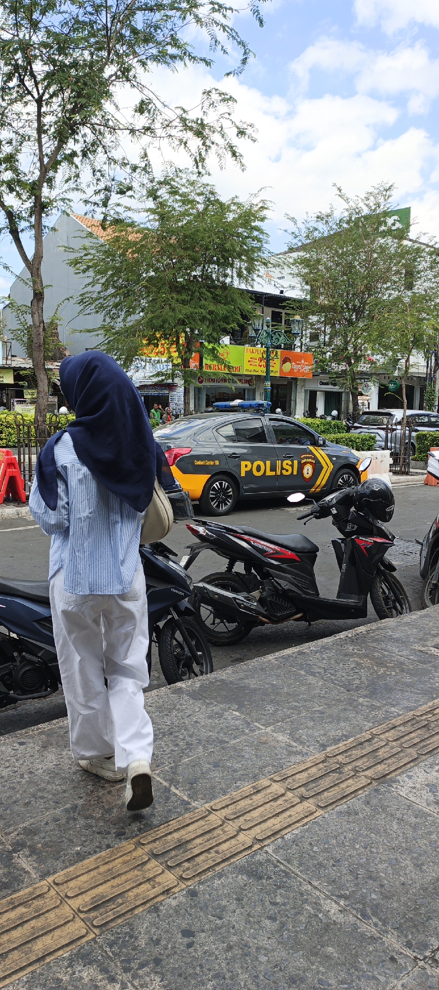 In Yogyakarta, Indonesia, a woman wearing a navy blue hijab and a light blue and white striped long-sleeved shirt is walking away from the camera. She has white pants and light-colored shoes. A beige bag is slung over her left shoulder. She is walking on a textured pathway with tactile paving for the visually impaired. Behind her, several parked motorcycles are visible, one of which is black with red accents. Further in the background, a dark gray police car with "POLISI" and yellow markings is parked on the street. The car has a blue and red light bar on its roof. Buildings with various storefronts and awnings are lined up behind the police car. Lush green trees with sparse foliage are scattered throughout the scene, and a cloudy blue sky is overhead. The overall setting appears to be a commercial or public area within the city.