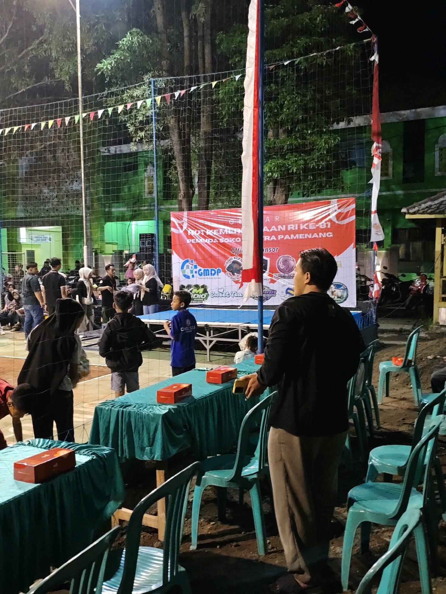 The image depicts an outdoor event at night, likely a volleyball match, in Pagu, Indonesia. A volleyball net is visible, and several people are gathered around a court, some watching the event and others in discussion. In the background, a large red banner with white lettering is displayed, and a green building can be seen. Two tables covered with teal cloths are set up, each holding red boxes. There are several teal plastic chairs scattered around the area. The lighting appears to be artificial, suggesting the event is taking place after dark. The presence of a banner with Indonesian text and the visual style of the buildings suggest a local community gathering.