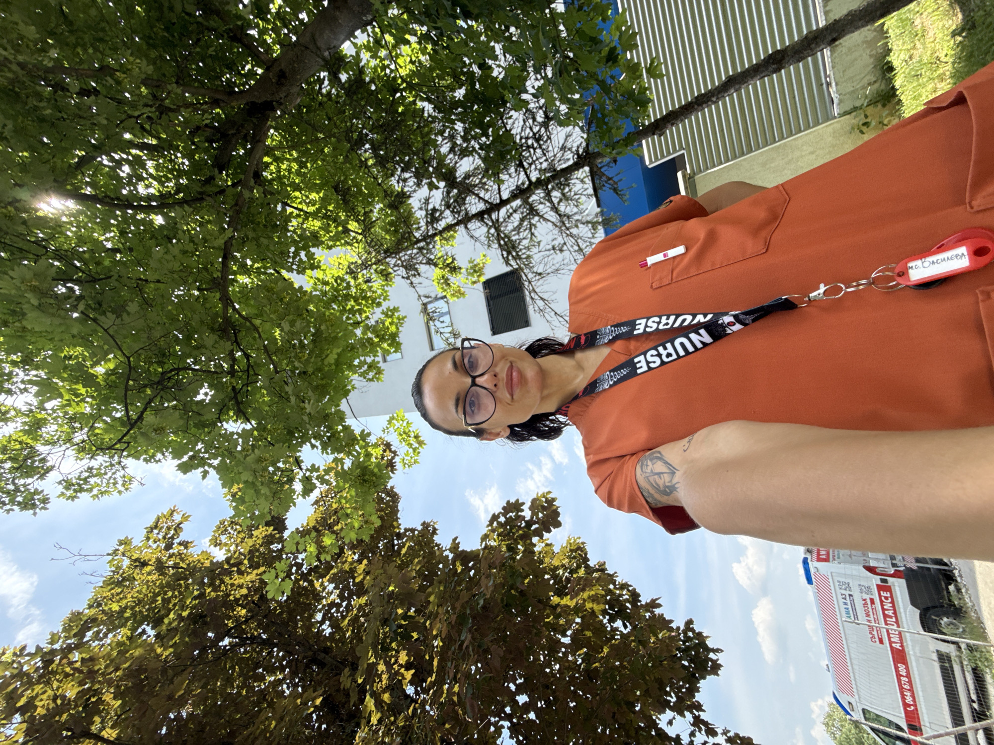 A female individual, identified as M.S. Vasileva (Medical Sister Vasileva) by the red key fob attached to her lanyard, is depicted in a low-angle selfie, positioned outdoors. She wears orange medical scrubs and black-framed glasses. A black lanyard with "NURSE" inscribed in white hangs around her neck, and a white and red pen is visible in her left chest pocket. A tattoo is present on her right bicep.
The background on the left reveals a white ambulance with red stripes, blue light bars, and the Bulgarian Cyrillic text "АМА И АЗ" and "СЪРЦЕ И МОЗЪК" (Heart and Brain), along with the English word "AMBULANCE" and the phone number "064/678 400." A fence runs behind the ambulance.
To the right, a multi-story white building with blue architectural elements and visible windows stands. The scene is heavily framed by trees with vibrant green and yellowish-green foliage, through which sunlight filters. The sky above is clear blue with sparse white clouds.
Based on the Bulgarian language elements, the "064" telephone area code specific to Pleven, and the "СЪРЦЕ И МОЗЪК" (Heart and Brain) branding on the ambulance, which identifies a major medical institution, the location is confirmed to be in **Pleven, Bulgaria**, specifically in the vicinity of the **UMBAL "Heart and Brain" Pleven hospital**.
