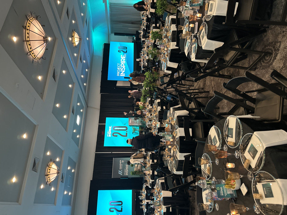 A conference room is set for an event, likely a banquet. Round tables with black tablecloths and transparent place settings are arranged in rows. Black folding chairs surround the tables. A stage with multiple screens displaying "Project Inspire 2.0" is visible at the front of the room. Several people are seated on the stage. The ceiling is decorated with ornate light fixtures and recessed lighting. The room appears to be well-lit.