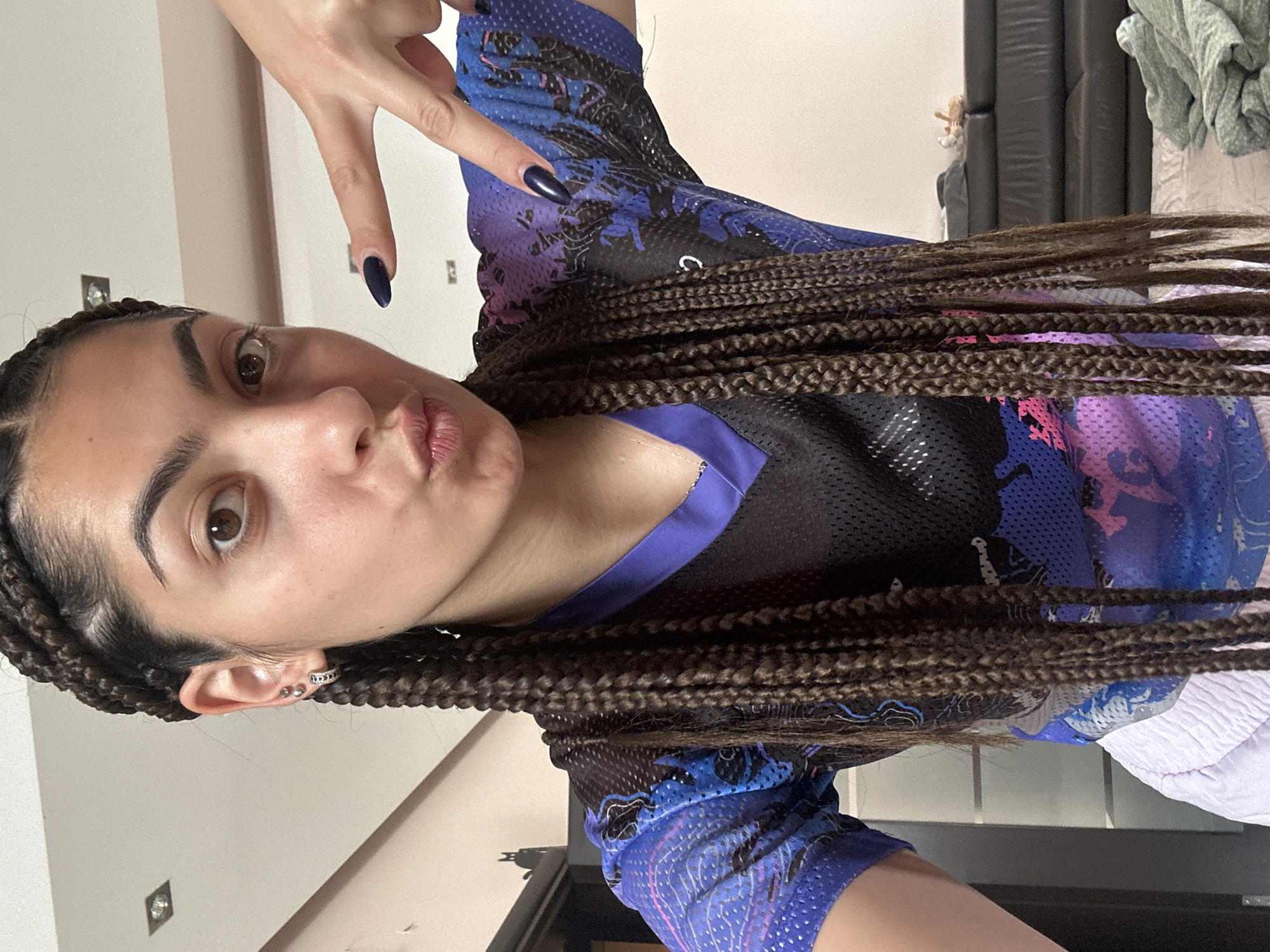 A person with long, braided dark hair and dark fingernails is shown in a close-up, vertical shot. The person is wearing a blue and purple patterned athletic top. They are making a peace sign with their right hand, with their index finger and middle finger extended. Their lips are pursed, and they are looking towards the camera. The background is blurry and appears to be indoors, with a wall and some furniture visible. The lighting suggests it could be daytime. There is no visible text in the image.