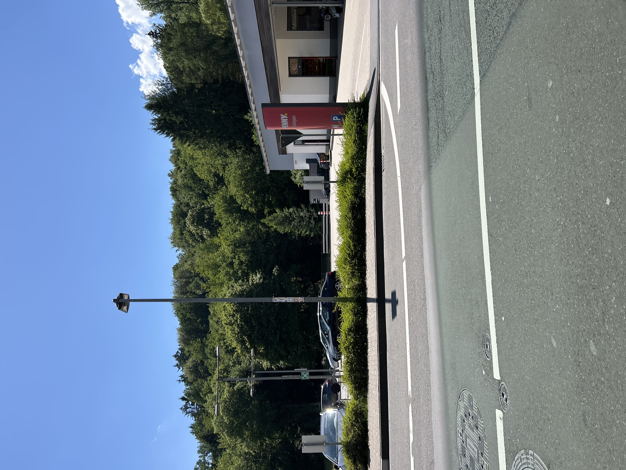 The image shows a street scene in Reichshof, Germany. The scene is a roadside with a building, trees, and a road. A red sign with the text "PENNY" and "en" is visible on the left side of the image, indicating a supermarket. There are several cars parked along the road and a street light. The sky is clear and blue, suggesting it is a sunny day. The image is taken from a slightly elevated perspective, looking down at the road and surrounding area. The overall mood is peaceful and ordinary, depicting a typical day in a German town.