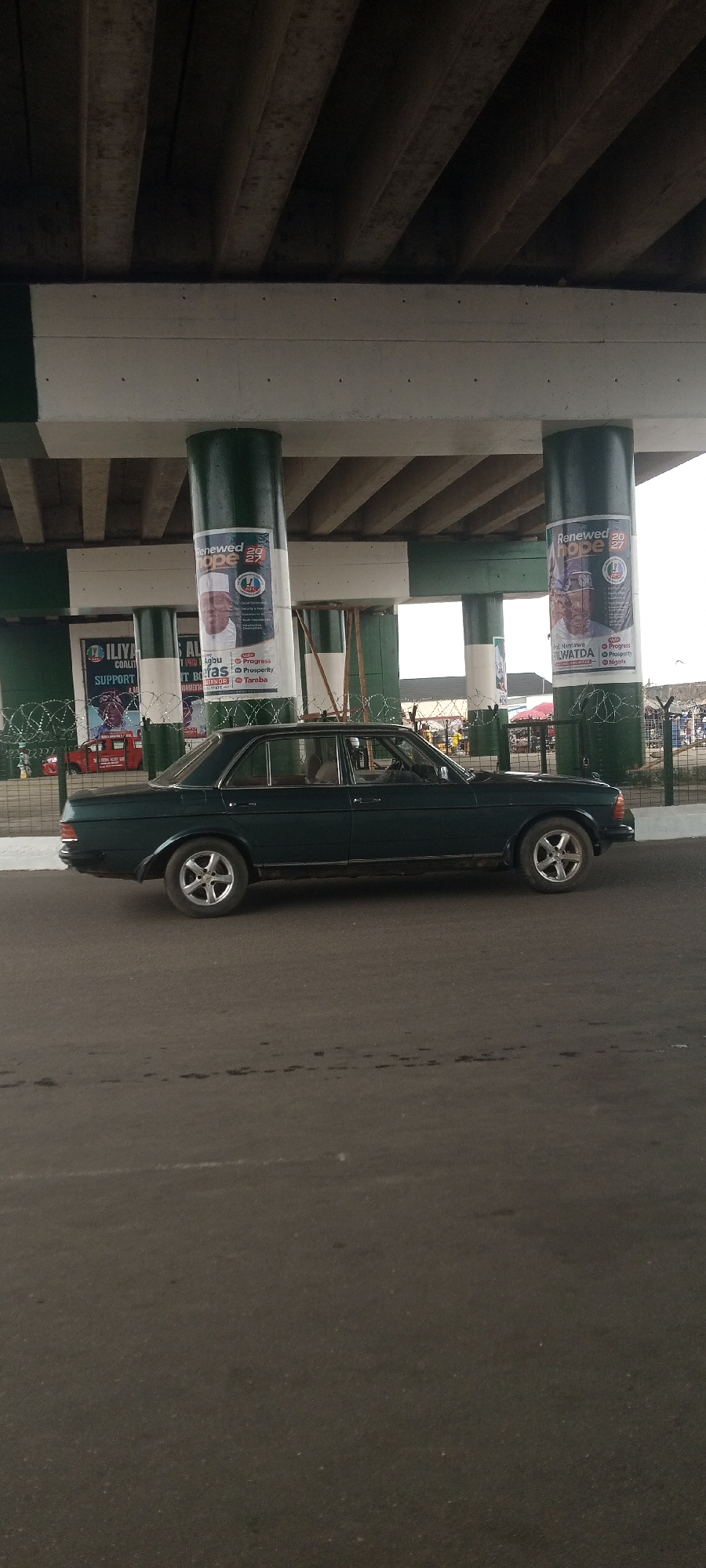 This media file captures a street scene in Jalingo, Nigeria, likely during daylight hours. The primary subject is a dark green sedan parked on a paved road beneath an elevated structure, possibly a bridge or overpass. The structure is supported by large, dark green cylindrical pillars.
On the pillars, several large posters are visible, featuring political campaign materials. One poster prominently displays the text "Renewed Hope 2027" and includes images of individuals. Another poster is for "ILIYA COALITION" and bears the word "SUPPORT". There is also text mentioning "Prof. Nentawe Yilwatda" with points like "Progress", "Prosperity", and "Nigeria". Barbed wire fencing is present behind the car and in front of some of the posters, suggesting a secured area or a boundary. In the background, there are hints of a busy environment with other vehicles, including a red commercial vehicle, and market stalls or shops. The overall setting appears to be an urban area with ongoing commercial activity. The weather appears to be overcast or hazy, as indicated by the diffuse light. The scene is static, with the car stationary and the overall impression being one of a typical day in a bustling Nigerian city.
--- OCR Start ---
Renewed 20
ope
Renewed 20
hope
COALIT
SUPPORT
AL
PON Agbu Progress
Berasto Prosperity
VERNOR Taraba
Nentawe
Progres
LWATDA Prosperty
Nigeria
--- OCR End ---