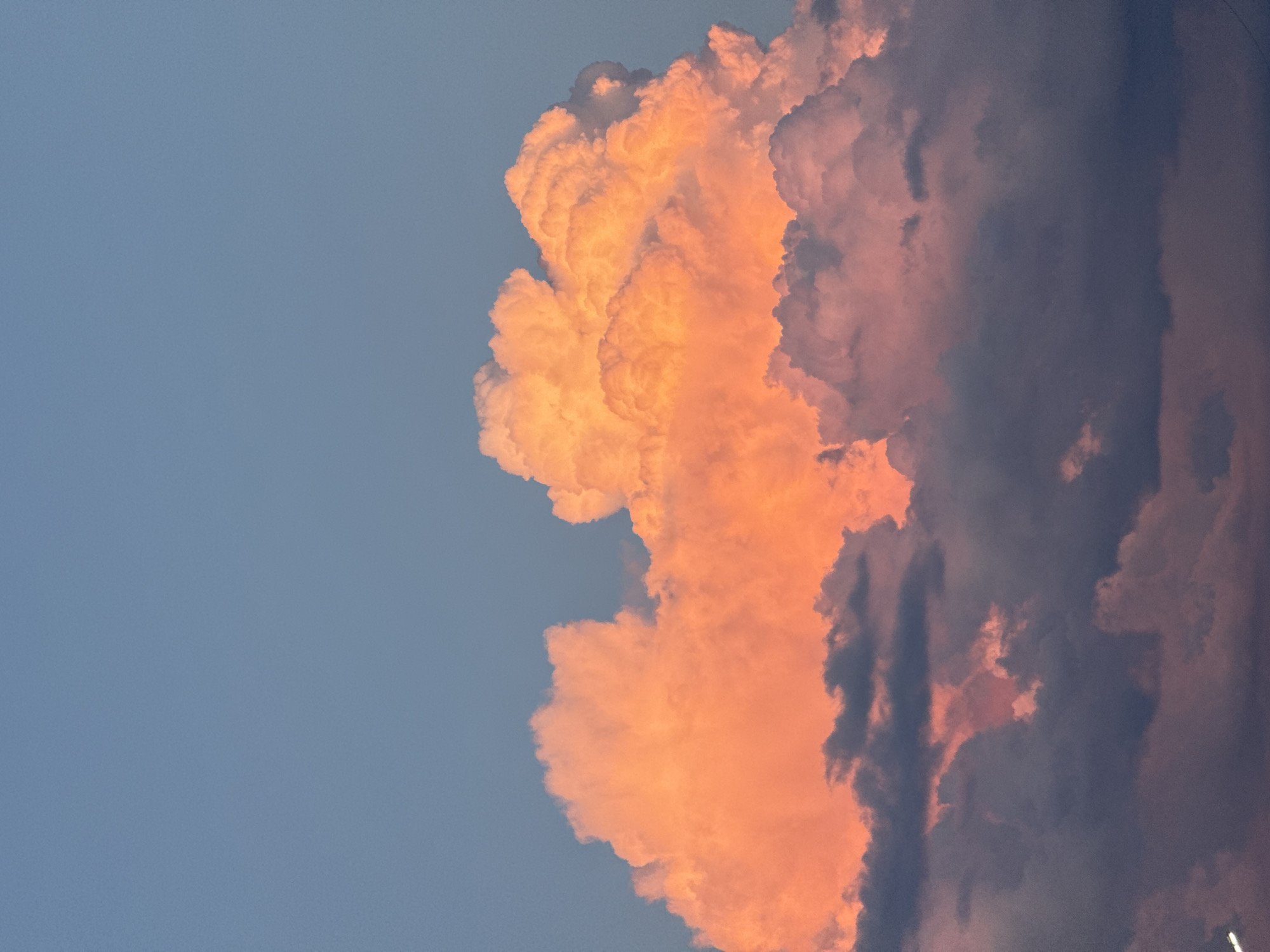 The image is a vertical, close-up shot of a dramatic sky filled with large, textured clouds. The clouds on the left are illuminated by a warm, golden-orange light, suggesting the presence of a sunset or sunrise. These clouds are voluminous and billowy, with sharp contours and bright highlights. To the right, the clouds are darker, appearing as deep shades of gray and purple, with some hints of pink and orange peeking through. This contrast between the brightly lit and shadowed clouds creates a sense of depth and dynamism. The background is a clear, pale blue sky. No people, objects, text, or specific location cues beyond the sky are visible. The overall mood is awe-inspiring and dramatic, capturing a powerful natural phenomenon. The time of day is clearly dusk or dawn, with the weather appearing to be partly cloudy with potentially developing storm elements suggested by the darker cloud formations.