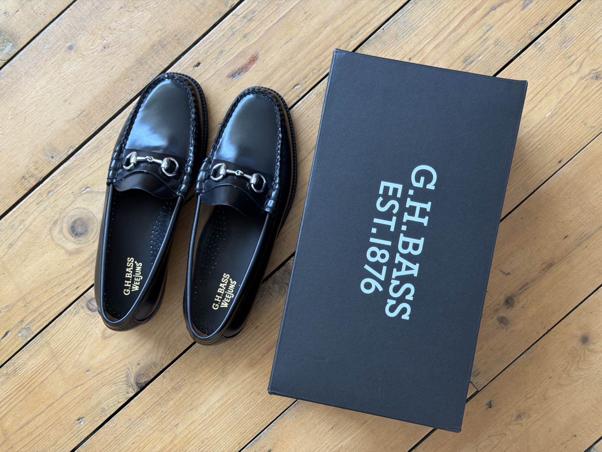 The image depicts a pair of black G.H. Bass Weejuns horsebit loafers lying next to a black G.H. Bass shoe box on a wooden floor. The shoes are positioned side-by-side, with their toes pointing slightly towards the upper left corner of the image. The box is located on the right side of the image, with the G.H. Bass logo and "EST. 1876" printed in white. The background consists of a light brown wooden floor, with visible grain and plank lines. The image appears to be taken from a high-angle perspective.
