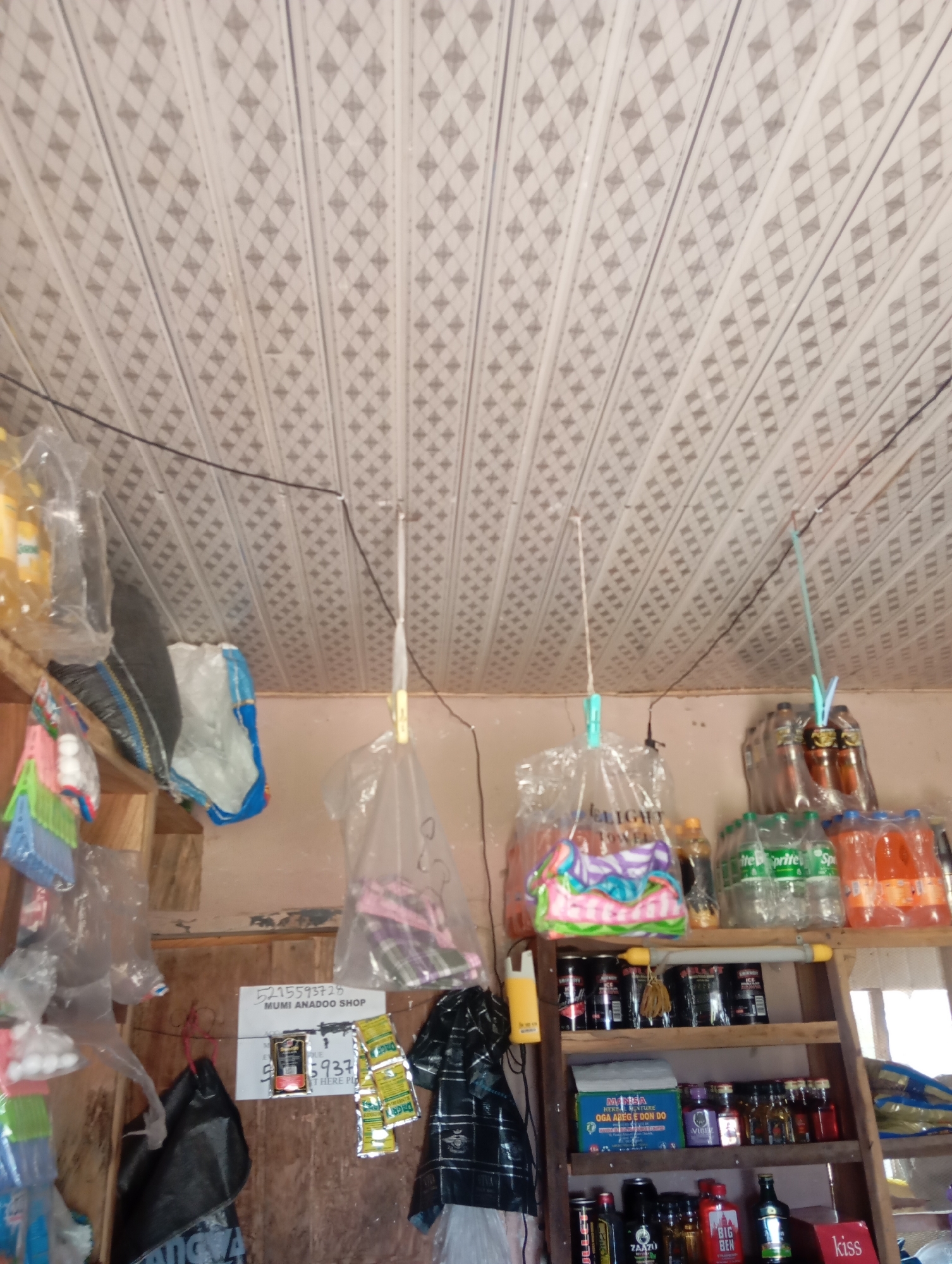 The image depicts the interior of a small shop in Jalingo, Nigeria. The ceiling is made of white and gray patterned panels. Various items are displayed, including plastic bags filled with products, bottles of soda, and other goods on shelves. A sign on the wall reads "MUMI ANADOO SHOP". The overall scene suggests a retail environment with a variety of consumer products.