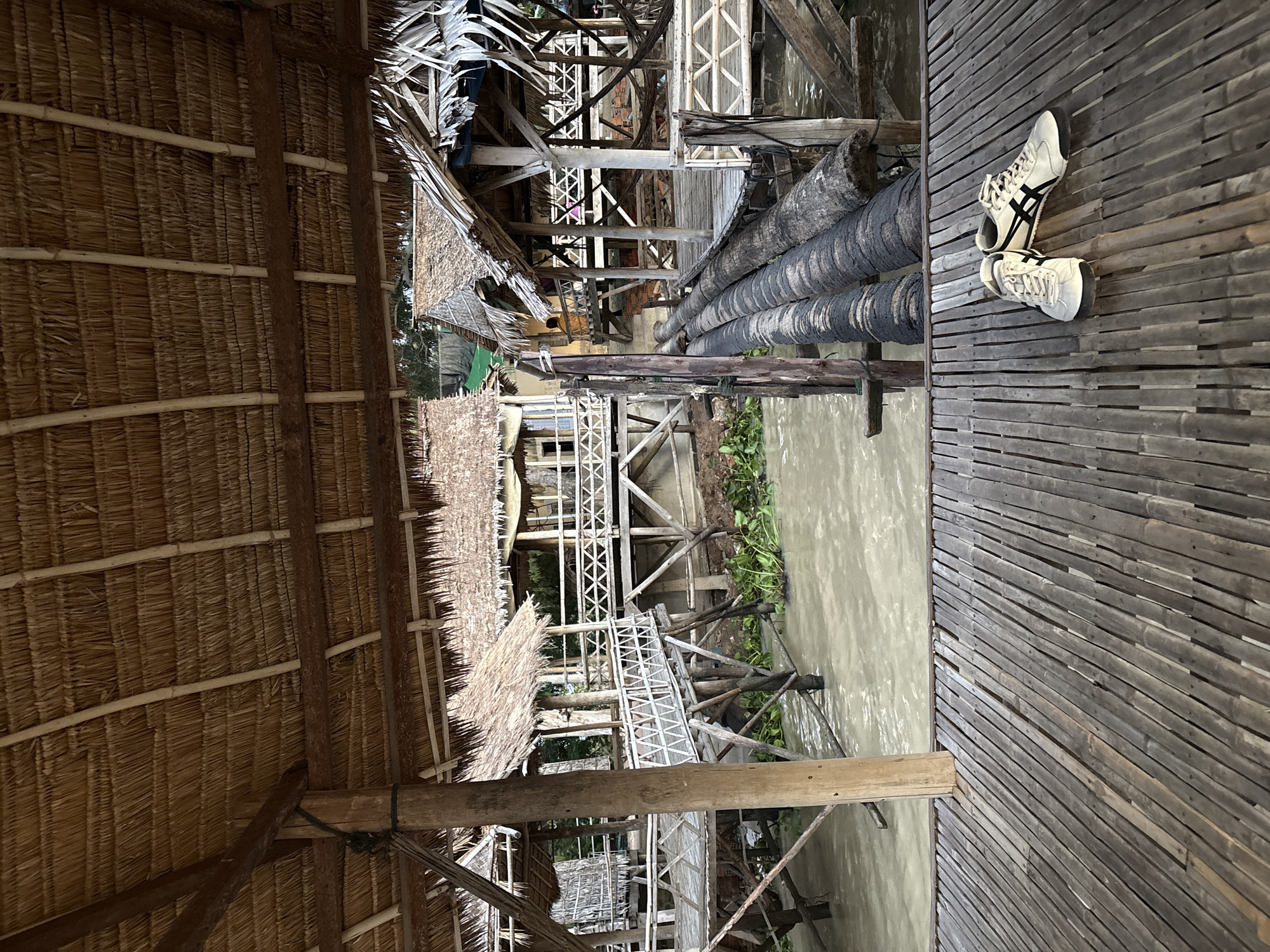 The image depicts a rustic waterfront scene, likely in Tonle Bati, Cambodia, characterized by thatched-roof structures and wooden walkways over murky water. In the foreground, a bamboo deck is visible, with a pair of white athletic shoes with black accents resting on it. Beyond the deck, a muddy river or canal is lined with stilted wooden buildings that also feature thatched roofs and open walkways. Some of these structures appear to be inhabited, with glimpses of activity or materials within. Aquatic plants float on the water's surface. The overall impression is one of a simple, possibly rural or tourist-oriented, environment on the water.
