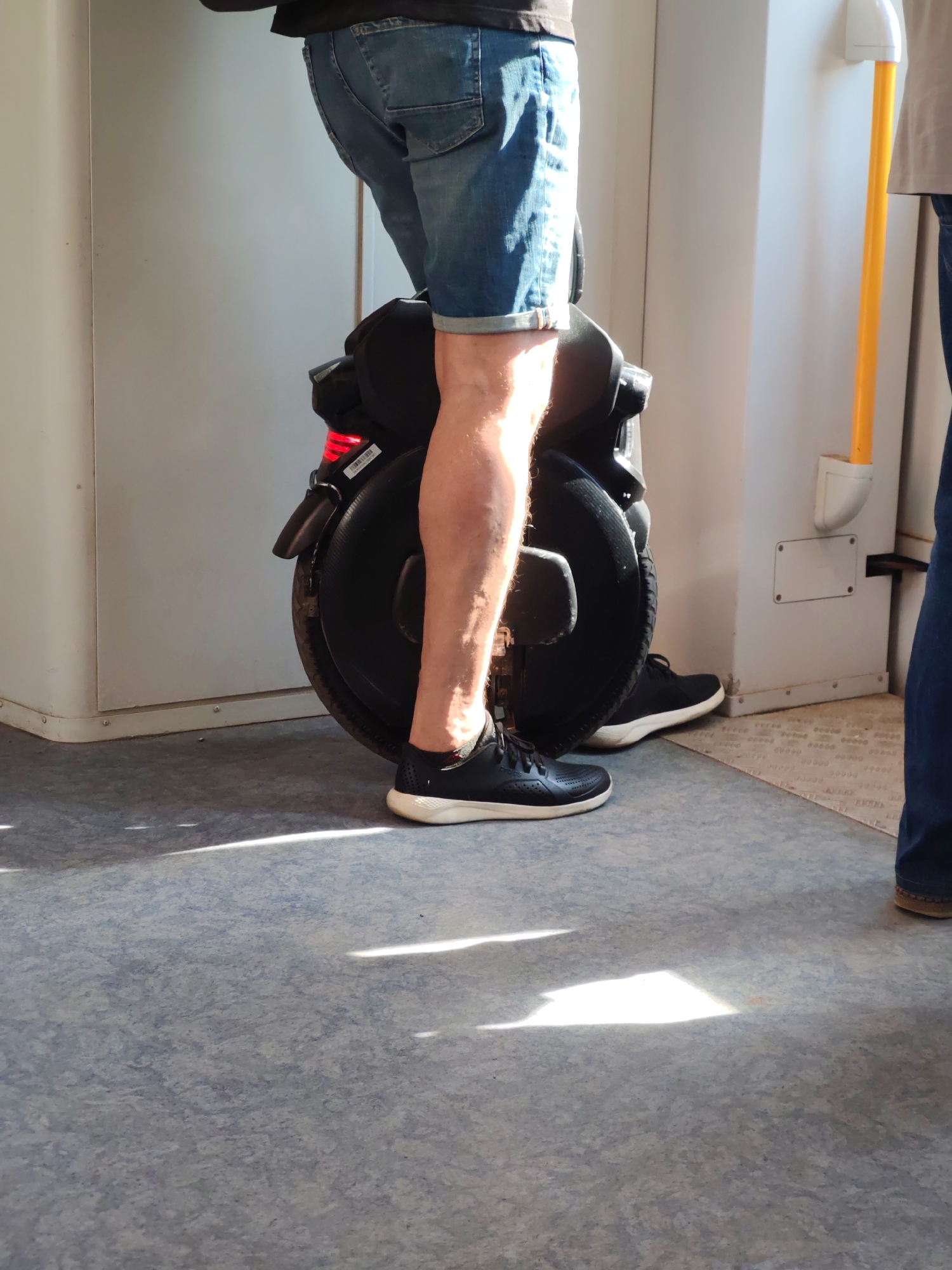 A person in denim shorts and black shoes is standing on a black electric unicycle inside what appears to be a train car. The person's right leg is visible, positioned to balance on the unicycle. Another person wearing jeans is partially visible on the right side of the image. The train car's interior includes a white wall, a yellow handrail, and a patterned floor. There are sunbeams visible on the floor.
It is not possible to confirm the location as Moscow, Russia, based on this image alone.