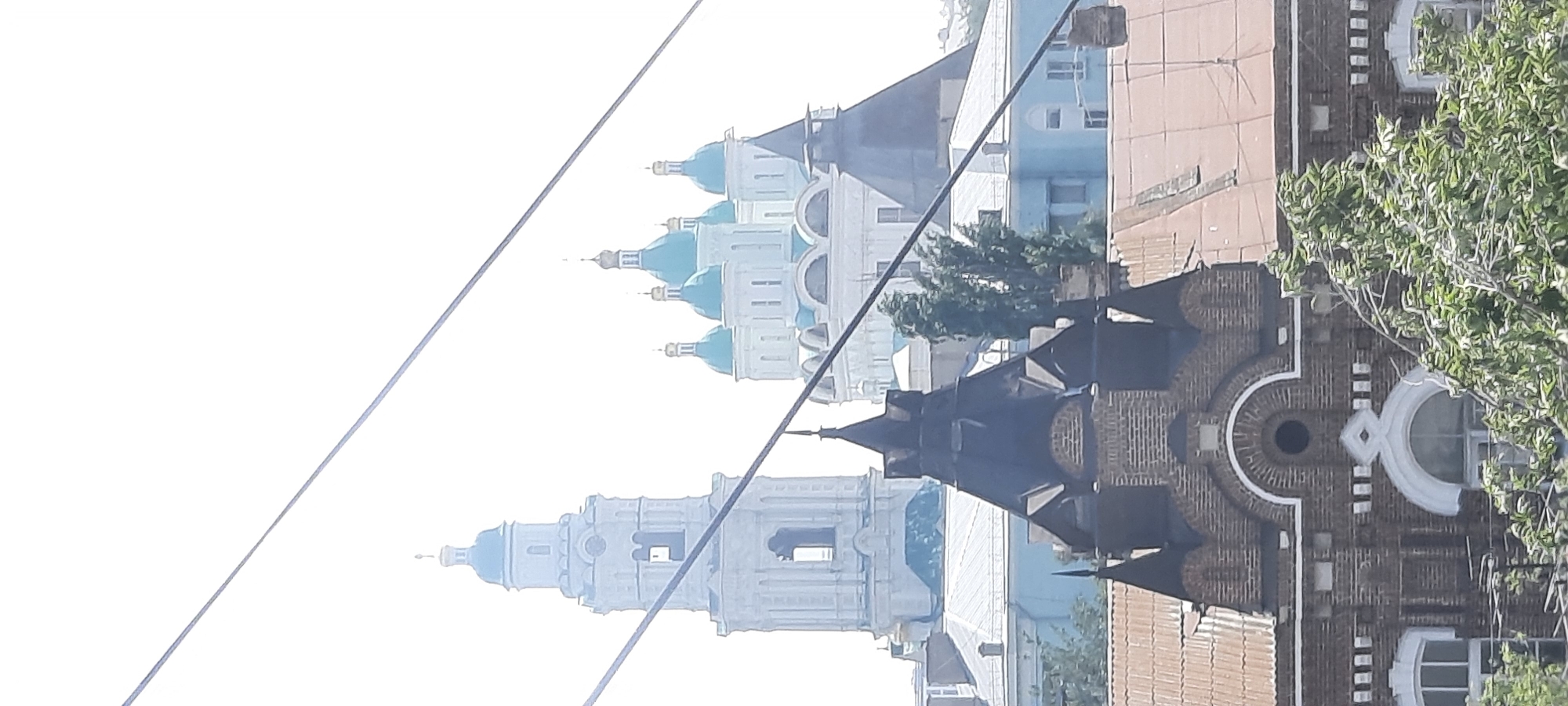 This is a photograph of a cityscape in Astrakhan, Russia, taken from an elevated viewpoint looking towards several prominent religious buildings. In the foreground, a dark, multi-gabled building with brickwork and a pointed, dark roof structure is visible, partially obscured by green foliage. Behind this building, a large, light-colored complex with multiple blue domes and a tall, slender bell tower with a clock face dominates the mid-ground. This complex is identifiable as the Dormition Cathedral and its bell tower within the Astrakhan Kremlin. Further in the background, another lighter-colored structure with blue domes is partially visible. The sky is bright and overcast, and several taut wires crisscross the frame, suggesting the photograph was taken near a power line or tramway. The overall scene depicts an architectural ensemble typical of historic Russian cities, with a blend of religious and secular structures.