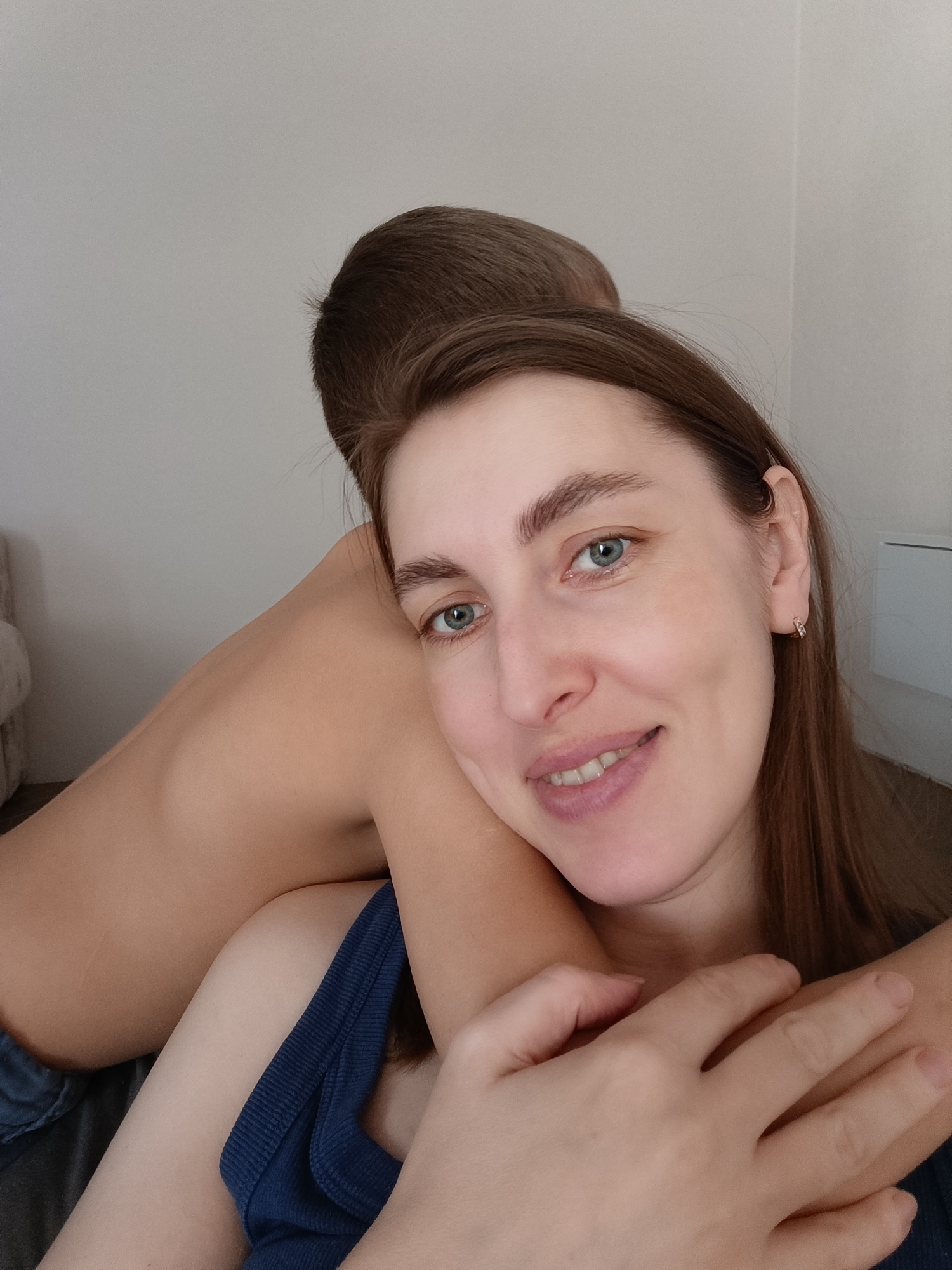 A woman with long brown hair and blue eyes is smiling into the camera. She's wearing a blue tank top and is being embraced by a person with short brown hair and bare skin, likely on their back. The location is indoors, possibly a home, with a plain white wall in the background. The image was likely taken in a city of Chaikovsky, Russia.