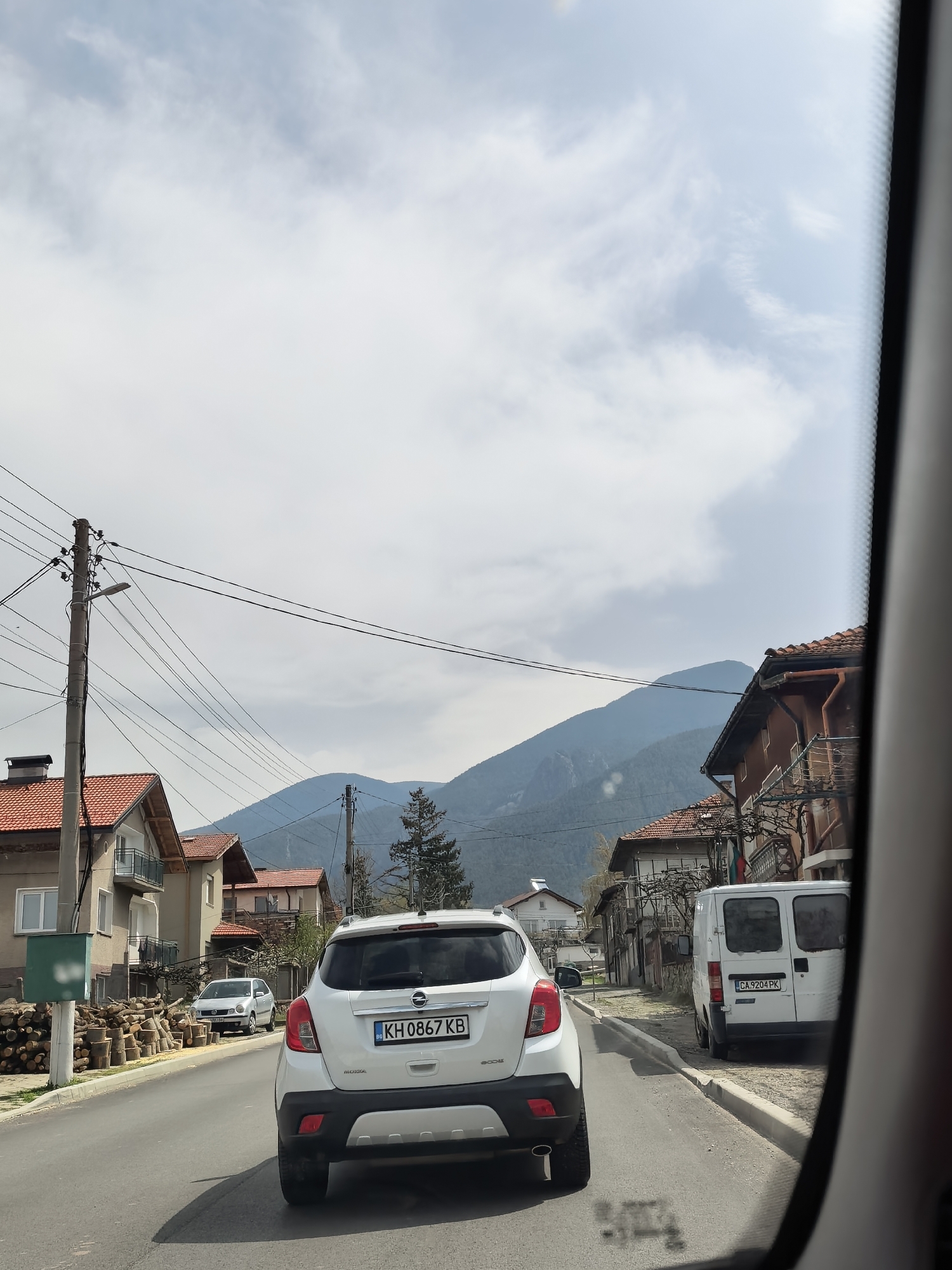A view from inside a vehicle, looking through the windshield at a street in a small town, presumably in Bulgaria based on the license plates. The road is asphalt and has a slight curve. In front, there is a white Opel Mokka with a Bulgarian license plate "KH 0867 KB". To the right, a white van with a license plate "CA 9204 PK" is parked. On the left side of the road, there are residential buildings with tiled roofs. There's a utility pole with wires running across the street. In the background, there are distant mountains under a cloudy sky. The angle indicates the image was taken from a vehicle. The overall environment appears to be a quiet residential area.