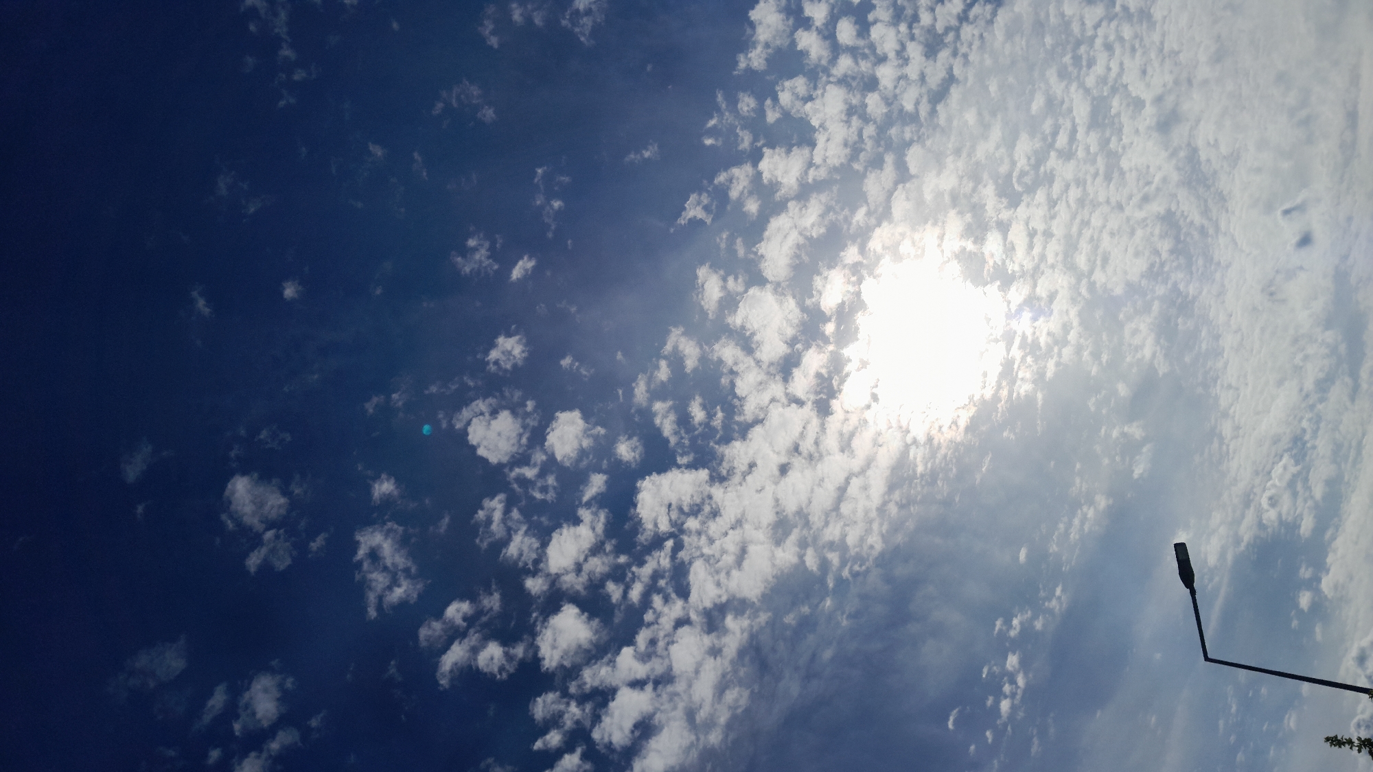 A streetlight with a dark pole and a single lamp fixture is silhouetted against a bright, midday sky filled with scattered white clouds. The sun is intensely bright and partially obscured by clouds, casting a hazy glow. The sky transitions from a deep blue at the top to a lighter blue near the horizon. This scene was captured in Krakow, Poland.