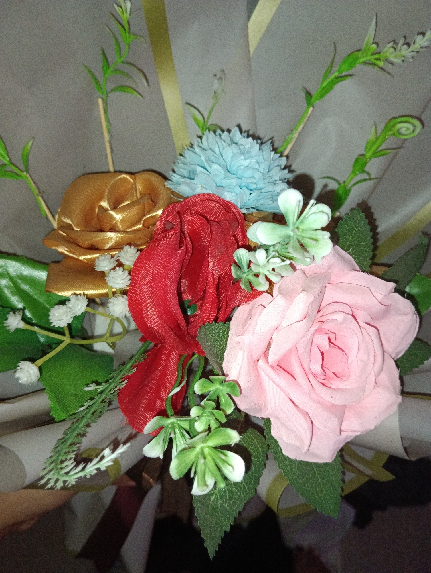 The image shows a close-up view of a bouquet of artificial flowers. The bouquet contains a large pink rose, a red rose, a gold rose, and a blue textured flower. There are also small white flowers and green leafy sprigs scattered throughout. The flowers are arranged in a way that suggests they are part of a gift or decoration. The background is a soft, neutral gray, possibly a wall or paper. The lighting is somewhat dim, casting subtle shadows on the petals and leaves. The arrangement is presented in a way that the flowers are the sole focus of the image. The image does not depict any specific location in Pematang Jaya, Indonesia, nor does it contain any people or actions.