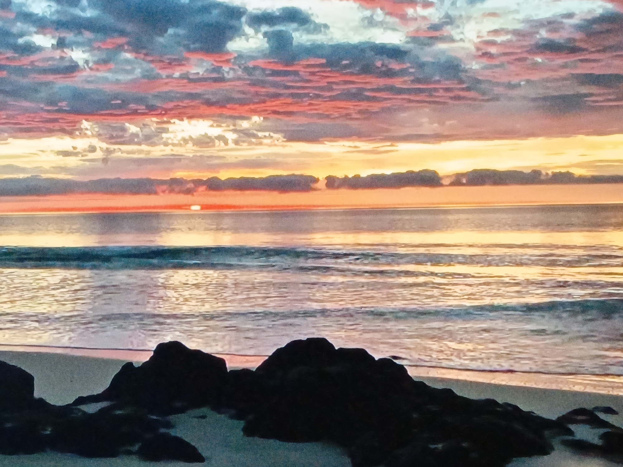 The image depicts a sunrise scene at a beach, likely Playa Caribe, near Juan Griego, Venezuela, known for its coastal views. The sky is filled with multi-colored clouds in shades of pink, orange, yellow, and gray. The sun is just above the horizon, reflecting a bright light across the water. Waves gently roll towards the sandy beach. In the foreground, dark rock formations are scattered along the shoreline. No people are visible in the shot.
