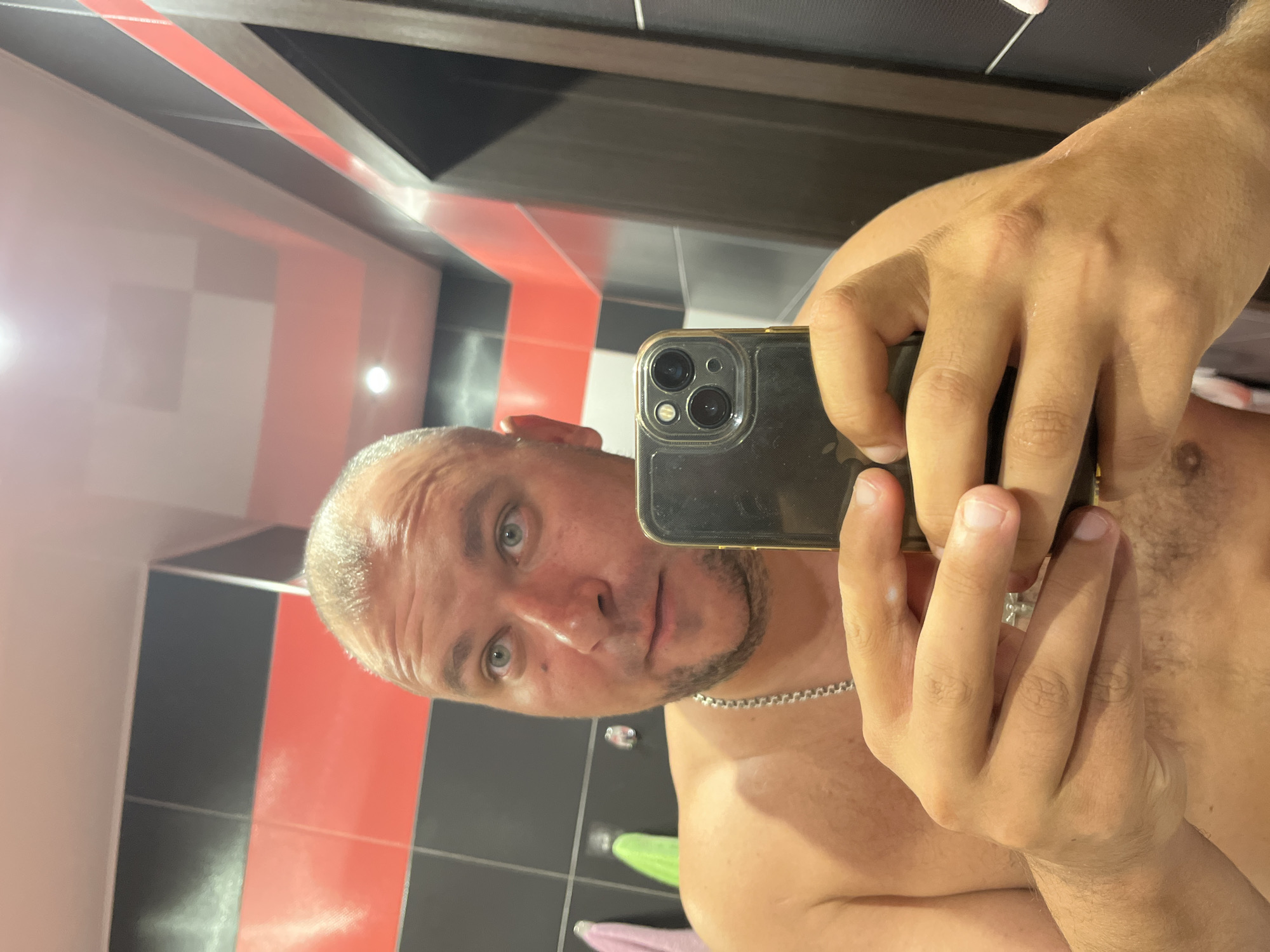 A man is captured in a mirror selfie indoors, appearing shirtless and wearing a silver chain necklace. He has short, light-colored hair, light eyes, and light facial stubble, looking directly at the camera with a neutral expression. He holds a smartphone, likely an iPhone with multiple camera lenses and a clear case, in his right hand, with his left hand supporting it from below. The background features tiled walls with a bold pattern of black, red, and white/light grey squares and stripes, typical of a modern bathroom or similar interior. Reflections of artificial light sources are visible on the shiny wall surfaces. Behind him, two out-of-focus towels, one green and one pink, are hanging on the wall. No text is visible in the image.