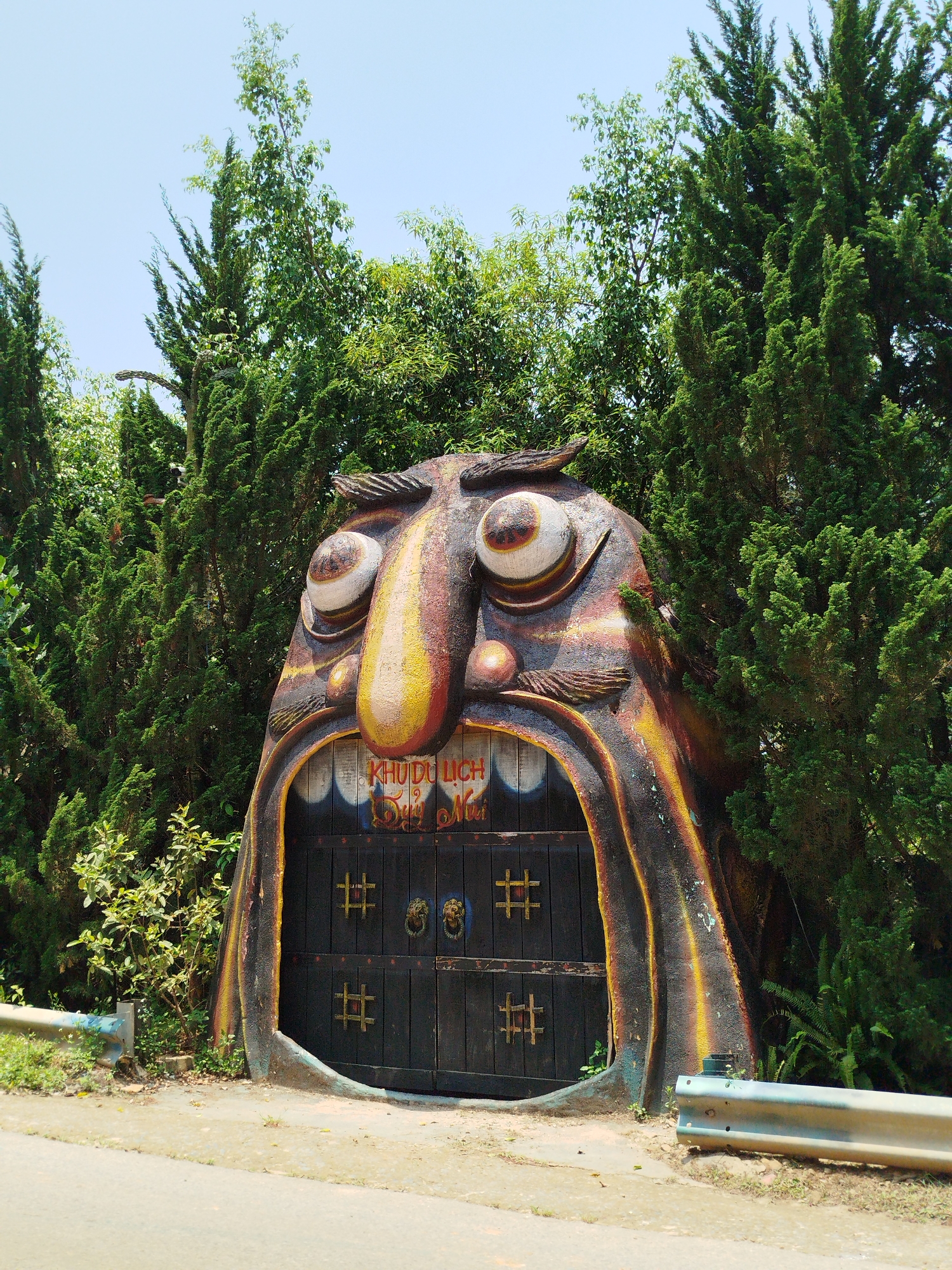 The image captures a striking, large-scale sculpture serving as an entrance gateway, set amidst lush greenery under a clear blue sky. The sculpture depicts a monstrous or stylized head with exaggerated features: wide, bulging eyes with distinct pupils, a large, bulbous nose, and an open mouth forming the archway for a gate. The sculpture is textured and painted in earthy tones of brown, yellow, and red, giving it a weathered, rustic, yet vibrant appearance.
Within the open mouth of the sculpture is a dark wooden gate, composed of vertical planks. This gate is adorned with square, grid-like metal patterns (resembling hash symbols) on its lower panels and two round, ornate metal elements, possibly lion head knockers, on the upper panels. Above the gate, inscribed on the inner arch of the sculpture's mouth, is text in Vietnamese. The upper line clearly reads "KHU DU LỊCH," meaning "TOURIST AREA." Below it, another line of text is partially visible, starting with "Suối" and followed by a less distinct word, possibly "Mơ."
The sculpture is flanked and surrounded by dense foliage. Tall, slender evergreen trees, likely conifers, create a thick backdrop and frame the entrance. Some broader-leaved green trees are also visible, particularly towards the top and behind the evergreens. A narrow road or path runs in the foreground, with a metal guardrail visible on both the left and right sides, indicating proximity to a roadside. The scene is brightly lit by natural sunlight, suggesting a clear day, likely late morning or early afternoon, with minimal shadows. The overall impression is an inviting yet whimsical or fantastical entrance to an attraction, located in Păng Tiêng 1, Vietnam.