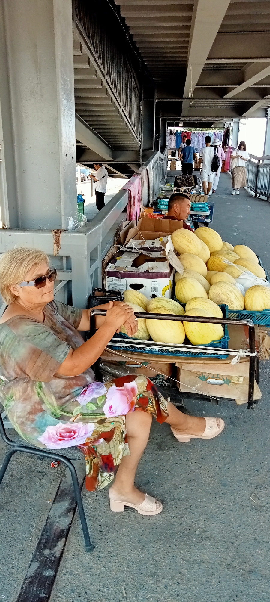 In Almaty, Kazakhstan, an outdoor market scene unfolds beneath a pedestrian overpass. The foreground features a woman with blonde hair and sunglasses, seated on a black chair. She wears a light-colored, patterned dress with large floral motifs. Her hands are clasped, resting on the armrest of a wheeled cart laden with a large quantity of light-yellow melons. Behind her, several other people are visible, some walking along the overpass, others near stalls displaying various items, including clothing. The environment suggests a bustling, informal marketplace setting.