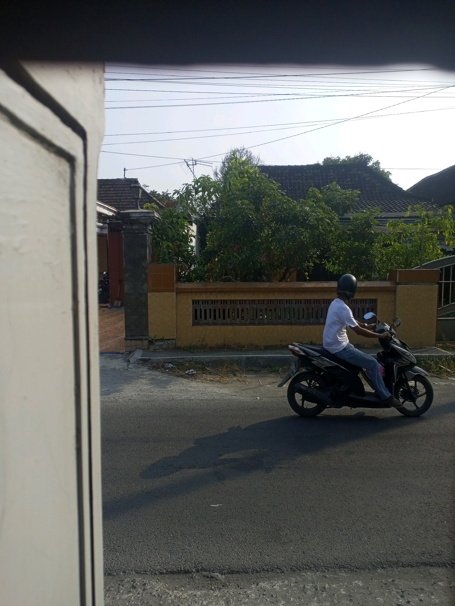 A person wearing a white t-shirt and blue jeans, with a black helmet, rides a black scooter down a paved street in Ngasem, Indonesia. The scooter is angled slightly towards the viewer's left. The person is holding onto the handlebars. Behind the scooter, a yellow and brown fence runs horizontally. Trees and the roofs of houses are visible beyond the fence. Power lines stretch across the sky. In the foreground, a portion of a light-colored structure, possibly a gate or wall, is out of focus and on the left side of the image. The sun casts a shadow of the scooter and rider onto the street.