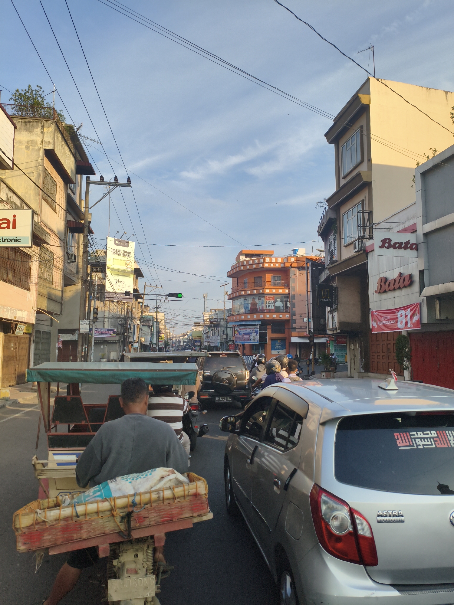 The image shows a street scene in the city of Tebing Tinggi, Indonesia, at dusk or dawn. The sky is a clear blue with wispy clouds, and the sun is casting a warm light on the buildings. The street is lined with multi-story buildings, some of which have businesses on the ground floor. There is a lot of electrical wiring crisscrossing overhead.

In the foreground, a person is riding a motorcycle-powered rickshaw. The rickshaw has a green canopy and a basket in the back filled with a white and blue item wrapped in plastic. The rider is wearing a grey shirt and is facing away from the camera.

Several vehicles are on the road, including cars and motorcycles. One silver Toyota car in the foreground has Arabic script on its rear window. The street appears to be busy with traffic. There are traffic lights visible, with a green light illuminated. Signs for businesses such as "Bata" and "Mosa" are visible on the buildings. The overall impression is of a typical urban street in Indonesia.