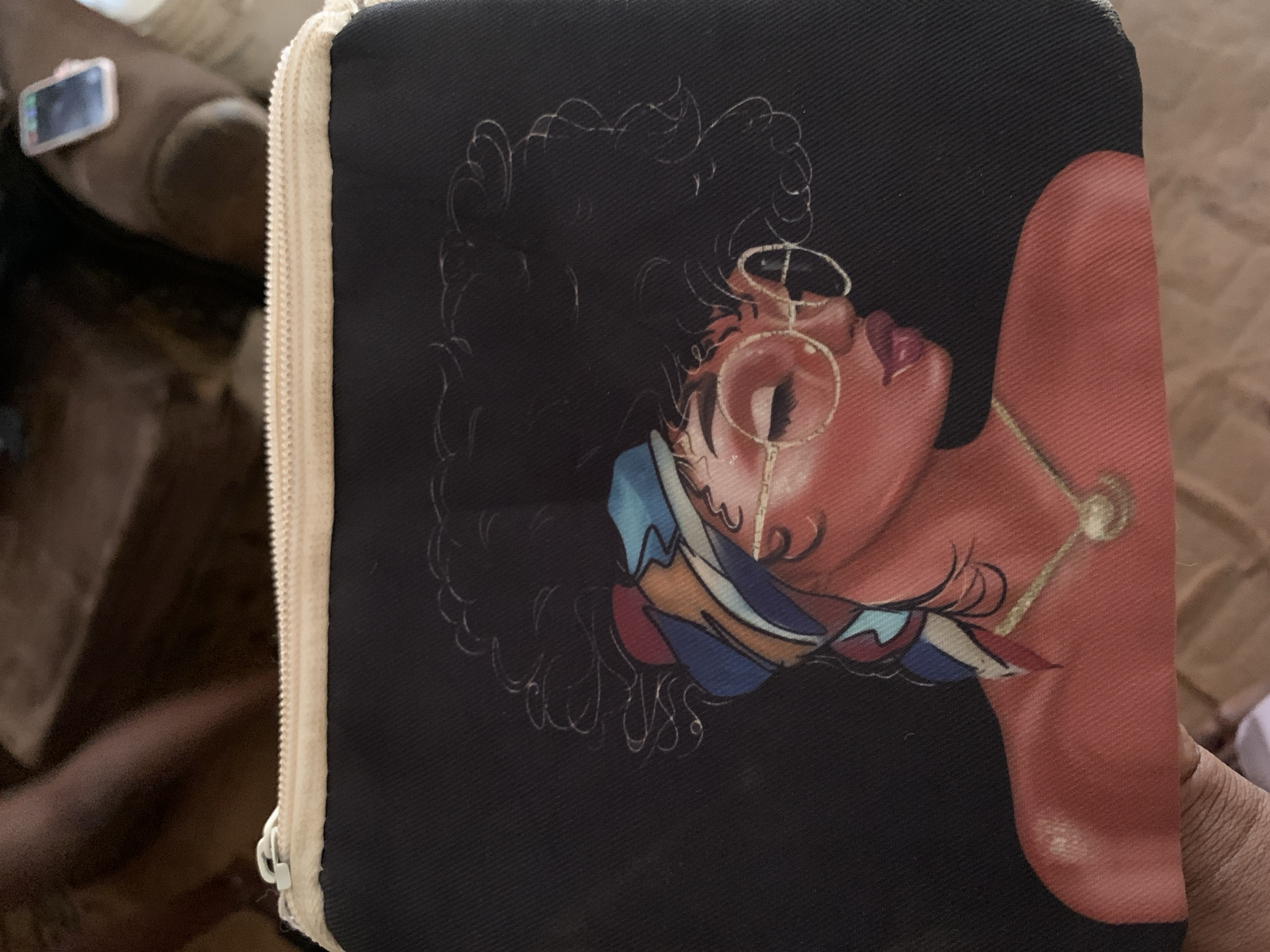 The image shows a close-up of a black cosmetic bag with a printed illustration of a woman with curly hair, wearing a colorful headscarf and round glasses. The woman has reddish-brown skin and is wearing a gold necklace. The bag is positioned on a brown surface, likely a table or a piece of furniture. In the background, a blurred human leg is visible, with a pink smartphone resting on it. The scene appears to be indoors. There is no specific information in the image that can be used to determine its location in Jos, Nigeria.
