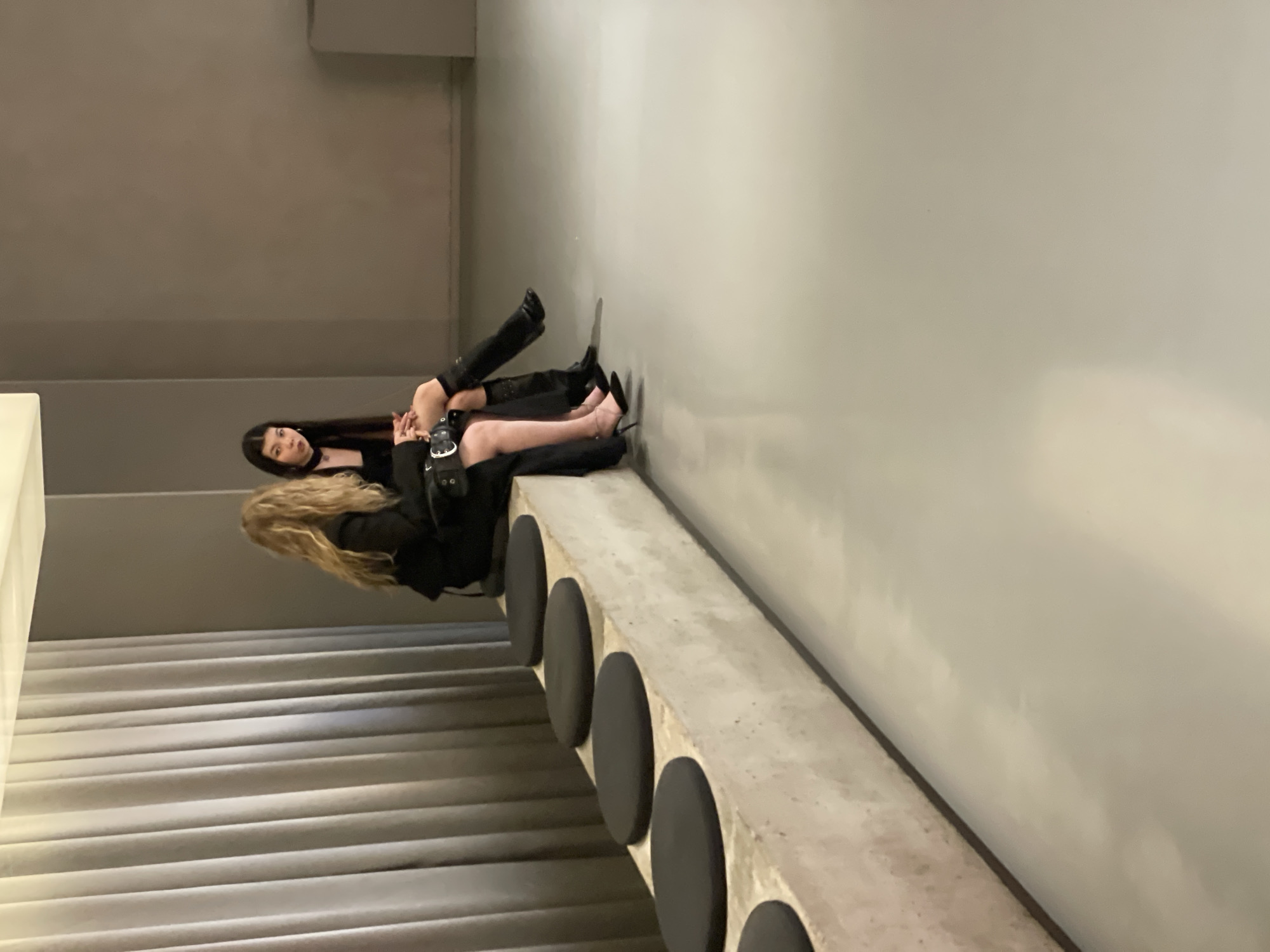 Two women are seated on a concrete bench in a modern, minimalist indoor setting. The woman on the left, with long dark hair and striking makeup, wears a black dress and tall black boots. She is leaning back, with one leg extended and the other bent. A small black bag is visible near her hands. The woman on the right has long blonde hair and wears a black jacket and sheer black tights with high-heeled shoes. She is posed with her legs crossed, resting against the wall. The background is a plain gray wall, and the bench is positioned alongside a set of grey, ribbed stairs. The lighting suggests an indoor environment, possibly during the day or evening. The overall atmosphere is stylish and contemporary, with a focus on the women's fashion and the minimalist architecture. There is no visible text in the image.