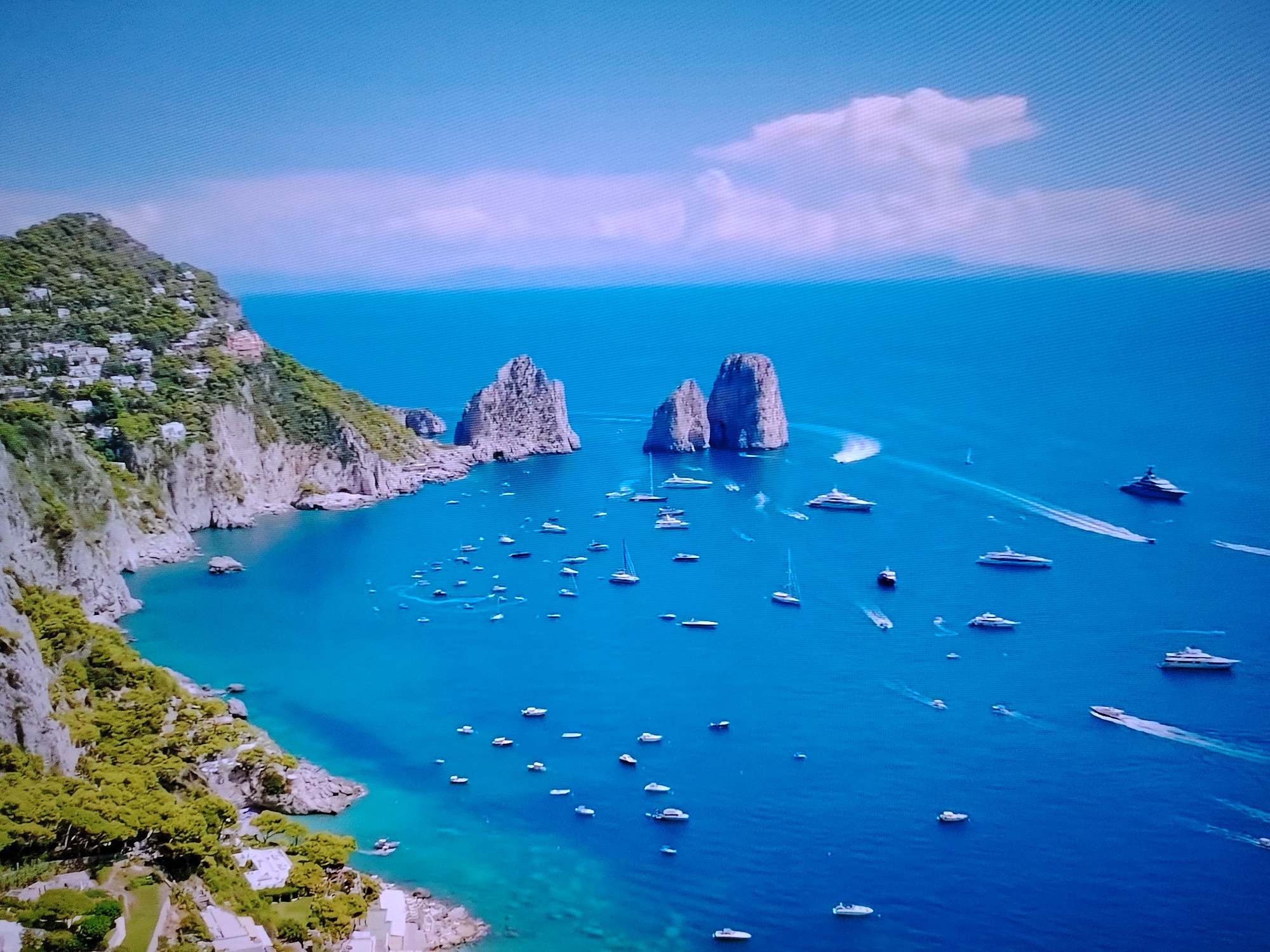 This is an aerial view of a coastal landscape. The scene features a rugged, green-covered cliff face with buildings nestled into the vegetation. The cliff drops dramatically into a bright blue sea filled with numerous boats of varying sizes, including yachts and smaller vessels. In the distance, prominent rock formations rise from the water, known as the Faraglioni. The sky is a clear blue with a few scattered white clouds. The image appears to be of the coast of Capri, Italy. This location is not in Huntingdonshire, United Kingdom.