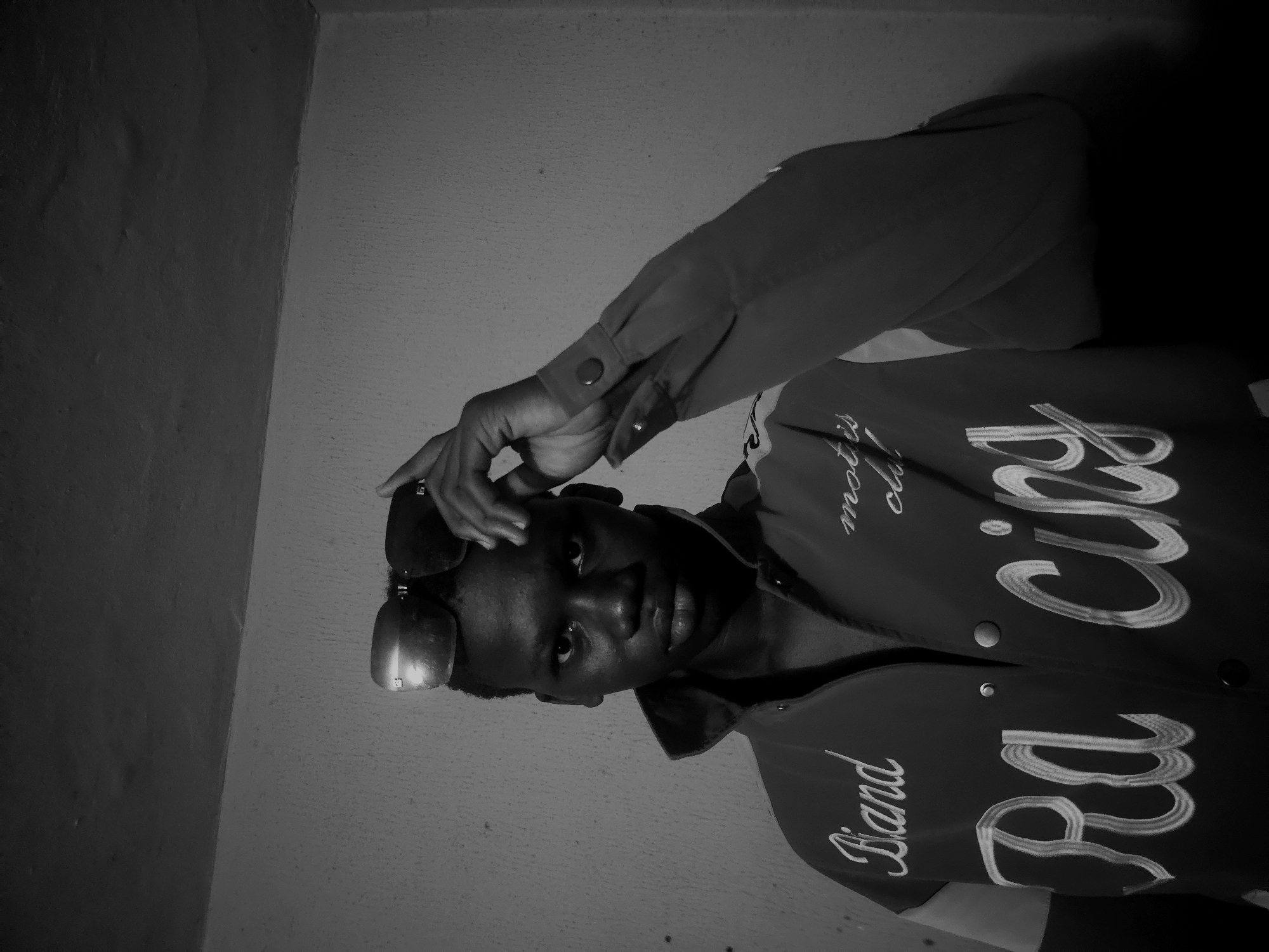 The black and white image displays a person, likely male, with a dark skin tone, posing against a plain, textured wall. The individual is positioned centrally, looking directly into the camera with a subtle, calm expression. They are wearing dark sunglasses pushed up onto their forehead, with their right hand gently resting near them.
The person is wearing a collared jacket, visible from the upper chest. The jacket has visible buttons down the front and on the cuffs of the sleeves. Several words are printed on the jacket in a light-colored, cursive script font. Clearly visible on the left side is the word "Band." Below it, the word "Red" is legible. Above "Red," the word "Club" is written in a similar script. Additionally, above "Club," a smaller, fainter script is visible, which appears to read "most it" or similar stylized text.
The background consists of a simple wall, showing a slight textural variation. The lighting is somewhat dim but even, contributing to the monochromatic aesthetic of the photograph. The setting appears to be indoors, possibly a room or hallway. The overall mood conveyed is casual and direct.