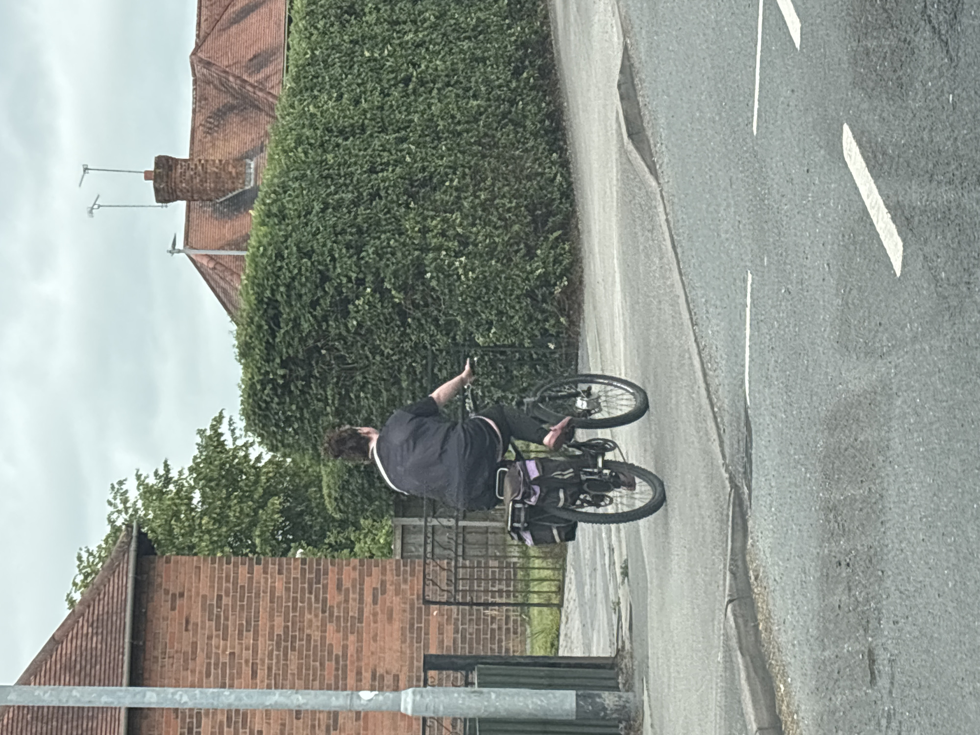 A person rides a bicycle with saddlebags on a paved sidewalk in Crewe, United Kingdom. The person is wearing a dark t-shirt and dark trousers, and their hair is dark and curly. The bicycle has a dark frame and black wheels. The background includes a brick building on the left, a dense green hedge in the center and right, and a residential roofline with chimneys and antennas in the distance. The sky is overcast. The road in the foreground has white markings.