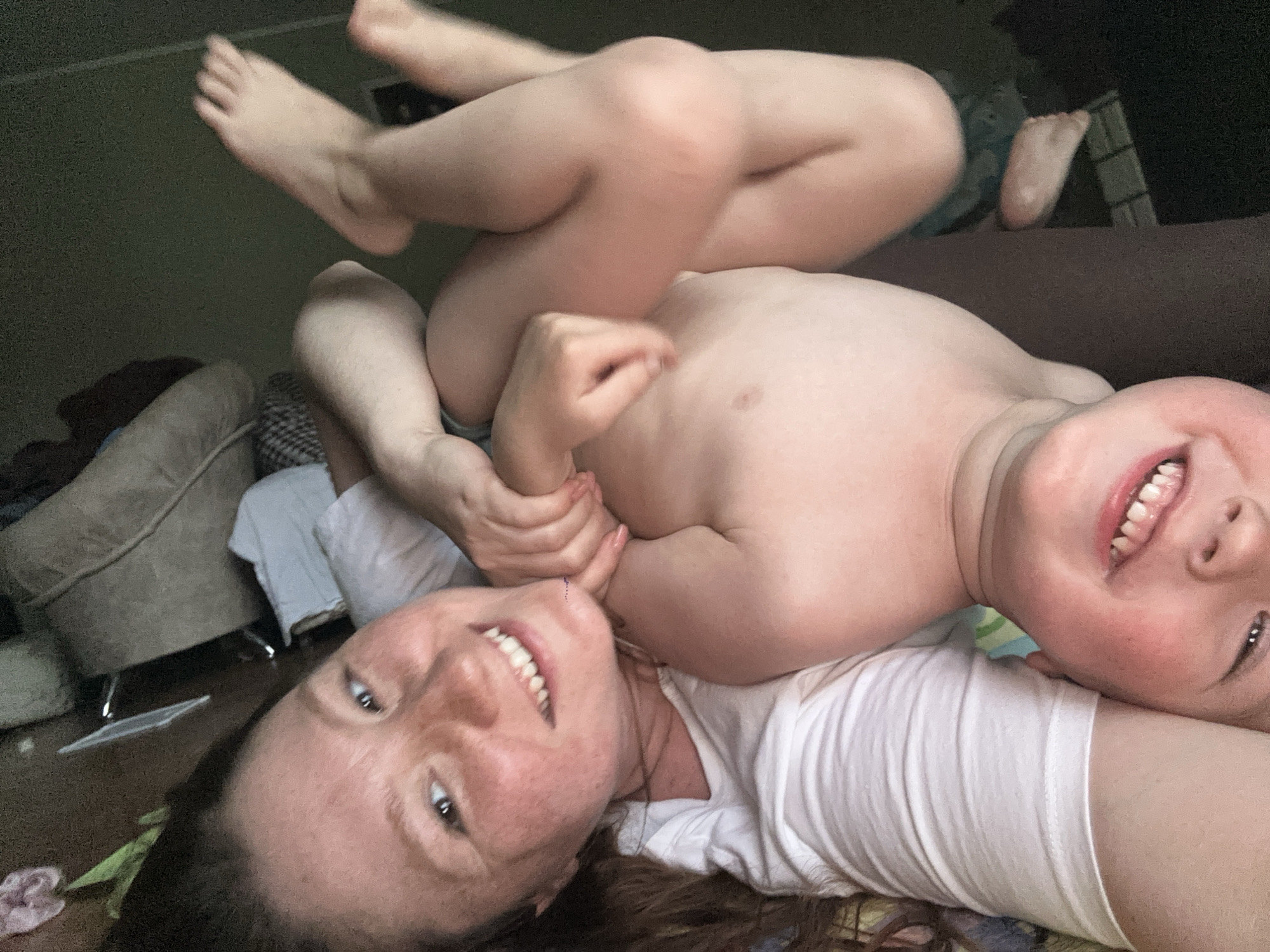 A woman lies on her back wearing a white t-shirt, with a young child lying on top of her. The child is shirtless. The woman and child are both smiling and looking at the camera. There is a sofa behind them, and what looks like a chair on the left of the image. Other bare feet are visible in the top left corner of the image.
I cannot determine whether the image was taken in Nizhny Novgorod, Russia.