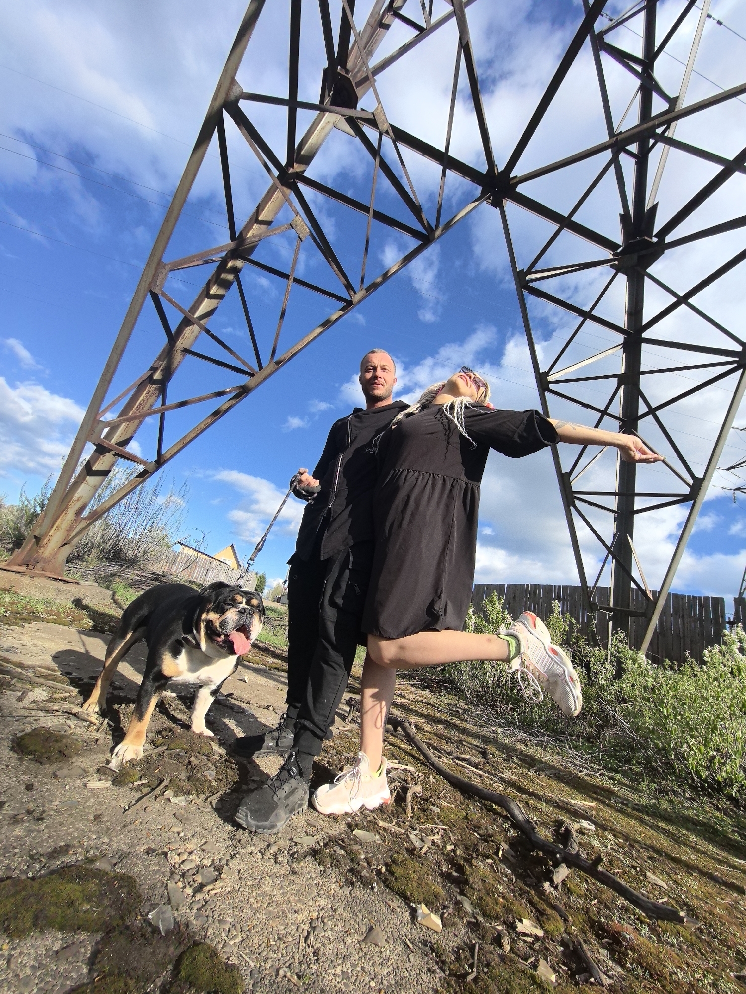 A wide-angle shot shows two people and a dog standing beneath a large metal electrical transmission tower. The location is outside the city of Noviy, Russia.
The man is on the left, dressed in black pants and a dark jacket, holding a leash connected to a black-and-tan bulldog. The dog is panting with its tongue sticking out.
The woman is on the right, wearing a black dress and white sneakers, with sunglasses on her head. She is posing with one leg raised and an arm outstretched.
The setting appears to be a slightly elevated, grassy area, with a dirt path and some vegetation. A wooden fence is visible behind the woman. The sky is blue with scattered clouds. The massive electrical transmission tower dominates the background.