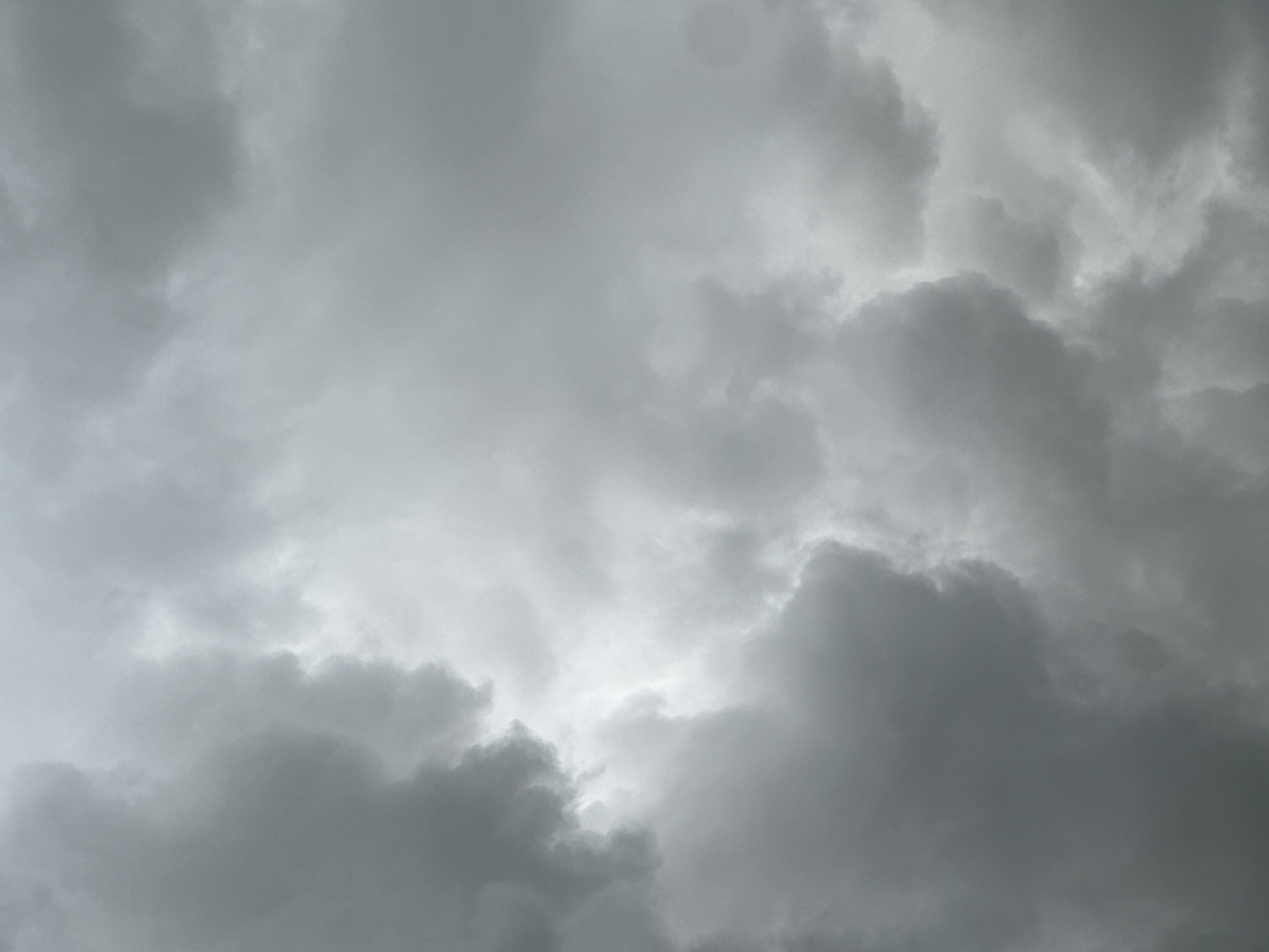 The media file displays a close-up view of an overcast sky filled with dense, gray clouds. There are no visible people, objects, or discernible activities taking place. The clouds vary in shade, with darker, more voluminous formations interspersed with lighter, almost translucent areas, particularly towards the center of the frame where a brighter, almost white patch suggests a slightly thinner cloud layer or a subtle light source breaking through. The overall impression is one of a heavily clouded, possibly gloomy or rainy day. There are no specific location cues or visible text. The time of day cannot be precisely determined but appears to be daylight given the general illumination, albeit muted by the thick cloud cover.
