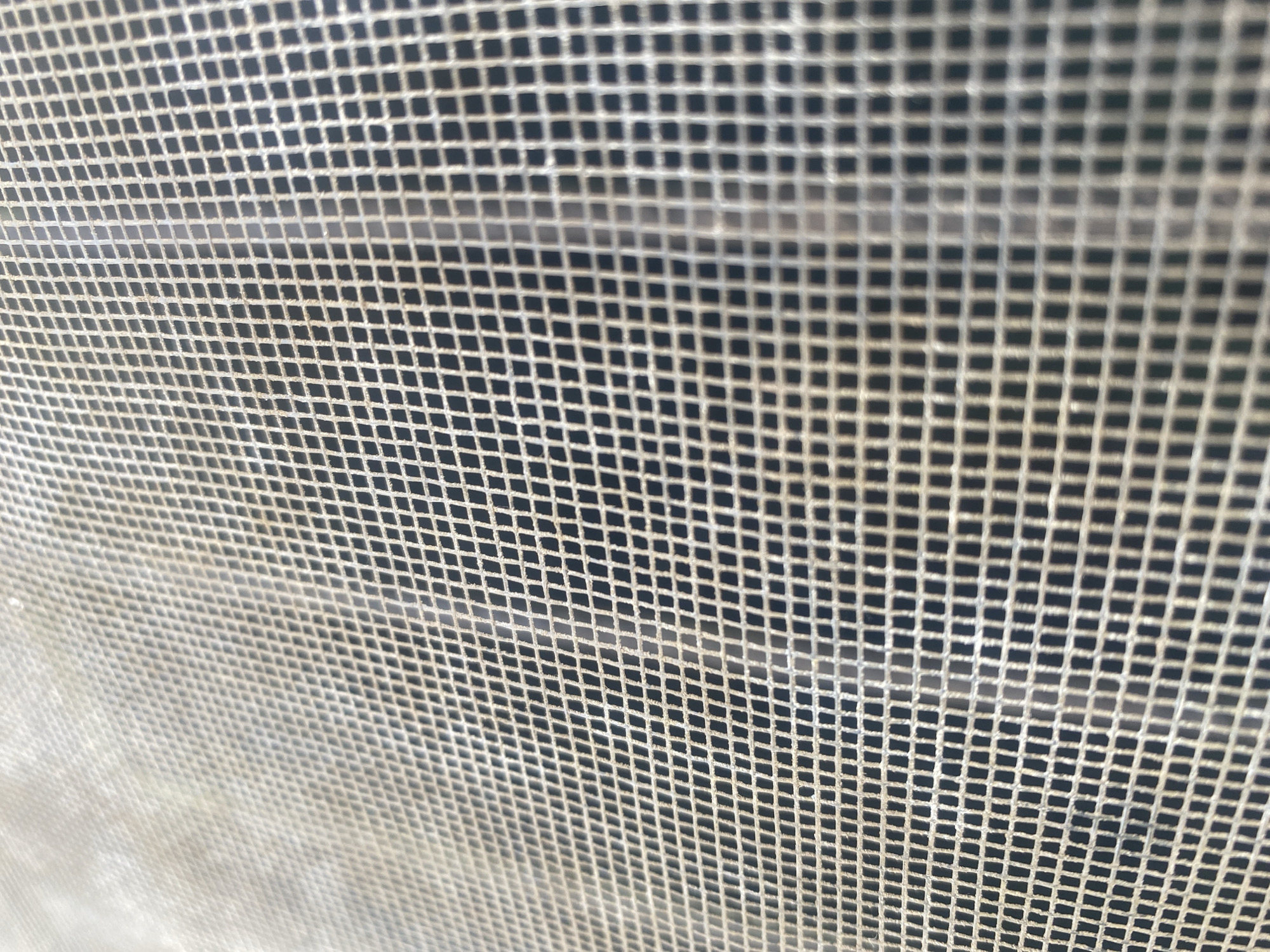 The image shows a close-up view of a window screen. The screen is made of a fine mesh material, creating a grid of small, square openings. The mesh appears slightly worn and dusty. The background seen through the screen is dark, suggesting either nighttime or a dimly lit interior. The location is indiscernible and unknown.