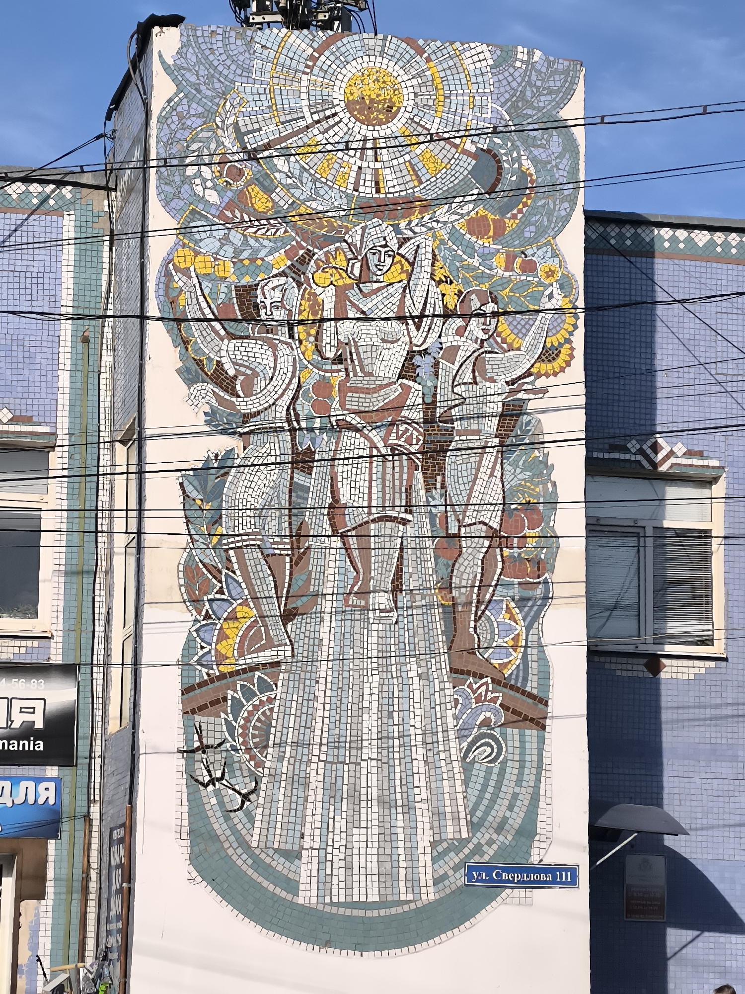 The image shows a large, colorful mosaic on the side of a building. The mosaic depicts three figures, possibly representing workers or mythological beings, holding up a sun-like orb at the top. The figures are stylized and are clad in patterned clothing. Surrounding them are abstract designs and floral motifs in shades of blue, green, yellow, brown, and white. The building itself appears to be made of lighter colored bricks, with some sections tiled in a light blue. Electrical wires are strung across the front of the mosaic. A street sign in Russian, reading "ул. Свердлова 111" (Sverdlov Street 111), is affixed to the building to the right of the mosaic, indicating the location is Sverdlov Street, number 111. The sky above is clear and blue. Part of a neighboring building, also tiled in blue, is visible to the right. Signs in Russian are also visible on the lower left part of the building.