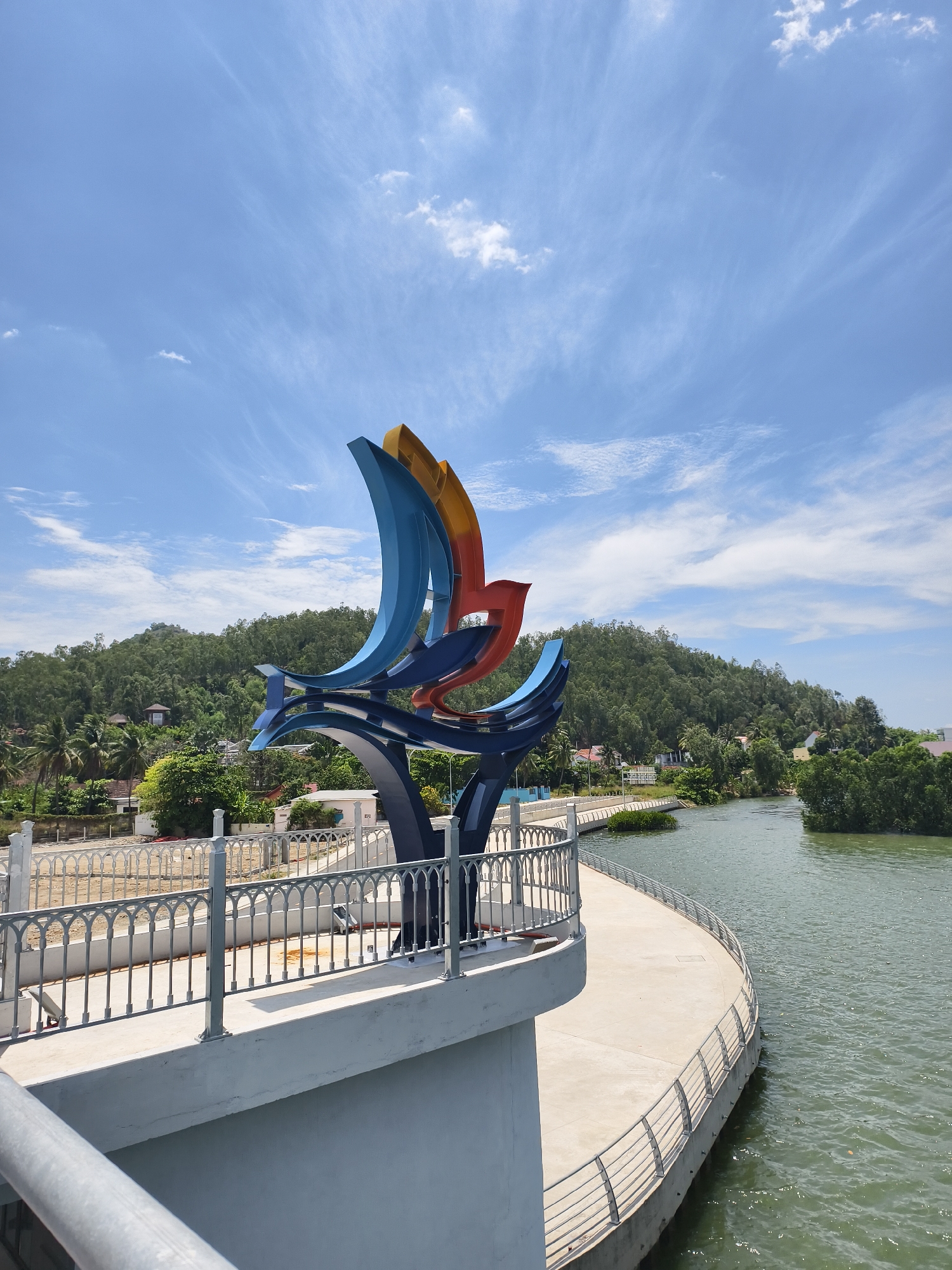 The image depicts a colorful public sculpture on a curved concrete walkway with railings, located in Tay Nha Trang Ward, Vietnam. The sculpture, primarily blue, with touches of orange and red, resembles abstract birds or sails. In the background, there is a green, tree-covered hill, and a body of water adjacent to the walkway, with an islet covered in trees. The sky is blue with scattered white clouds. A concrete wall with decorative railings is on the opposite side of the waterway.