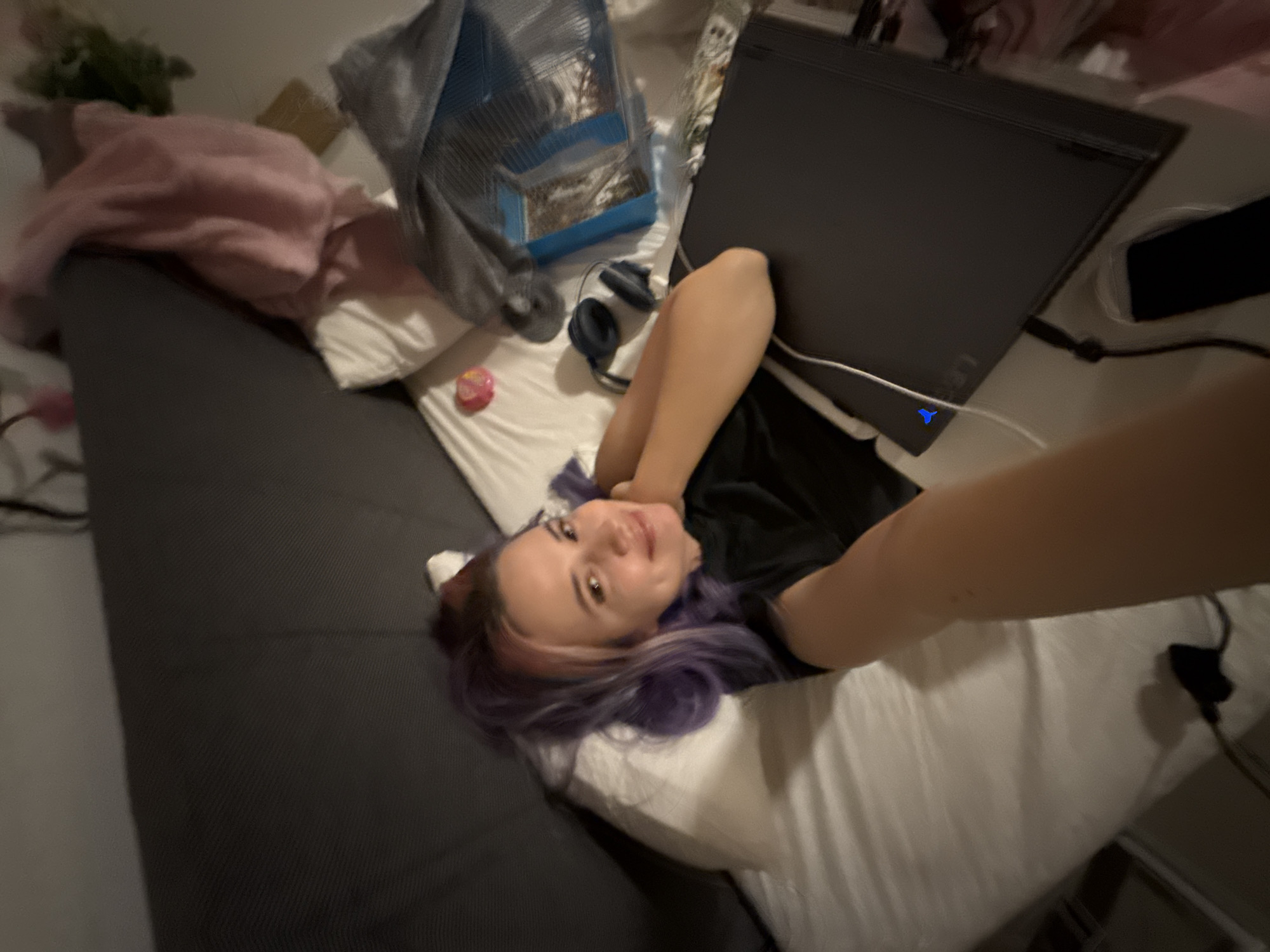 The scene depicts an interior domestic setting in the City of Zagreb, Croatia, likely a bedroom or living area. One adult female subject is visible, lying supine on a bed or couch, her head resting on a white pillow. She has light skin, and her hair is colored purple and reddish-brown. She is wearing a black t-shirt and is smiling towards the camera. Her right arm (from the viewer's perspective, left from the subject's perspective) is bent at the elbow and rests near her head, while her left arm (from the viewer's perspective, right from the subject's perspective) extends towards the camera, indicating she is holding the device taking the photograph.
Objects immediately around the subject include a dark-colored laptop positioned to her left (viewer's right) on the bed, with a white cable connected and a small blue light illuminated on its edge. A dark-colored smartphone is situated next to the laptop. A pair of dark blue over-ear headphones are on the white bed linens near her head. A small pink heart-shaped object rests on the bed near her right side (viewer's left). The background shows a dark gray headboard or backrest, crumpled pink fabric, a gray bag containing various items, and some green foliage, likely from a houseplant. The lighting in the scene is dim, suggesting evening or low-light conditions.