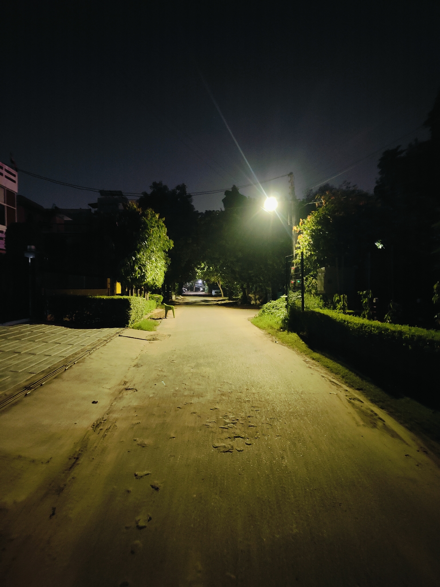 A long, paved path is illuminated by a streetlamp, casting a warm, yellowish glow. Trees and hedges line the sides of the path, creating a tunnel-like effect. The sky is dark, indicating that it is nighttime. The path is relatively empty, with a single green chair visible in the distance. The overall impression is one of a quiet, serene evening in a park or residential area. The specific location is a walkway in Gurgaon, India.