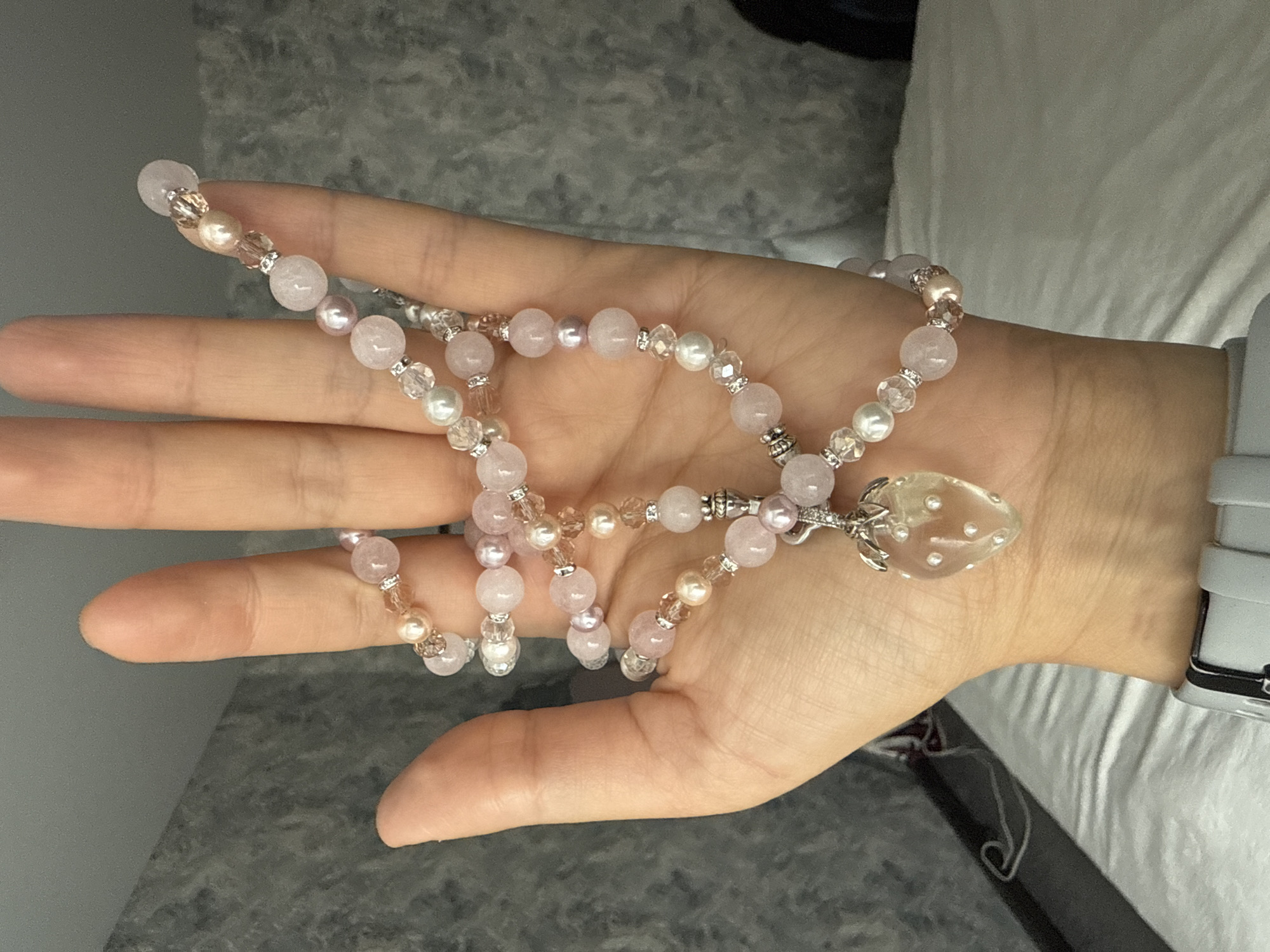 A person's left hand, with fingers slightly curled, is presented in the foreground, palm up. The hand is adorned with a delicate, multi-strand bracelet or necklace. The jewelry features a combination of light pink, pearlescent, and clear faceted beads. A translucent pendant, shaped like a strawberry with small white dot inclusions, hangs from one of the strands. The background is blurred, showing a textured grey wall and what appears to be a white duvet or bed. A grey smartwatch is visible on the person's wrist.