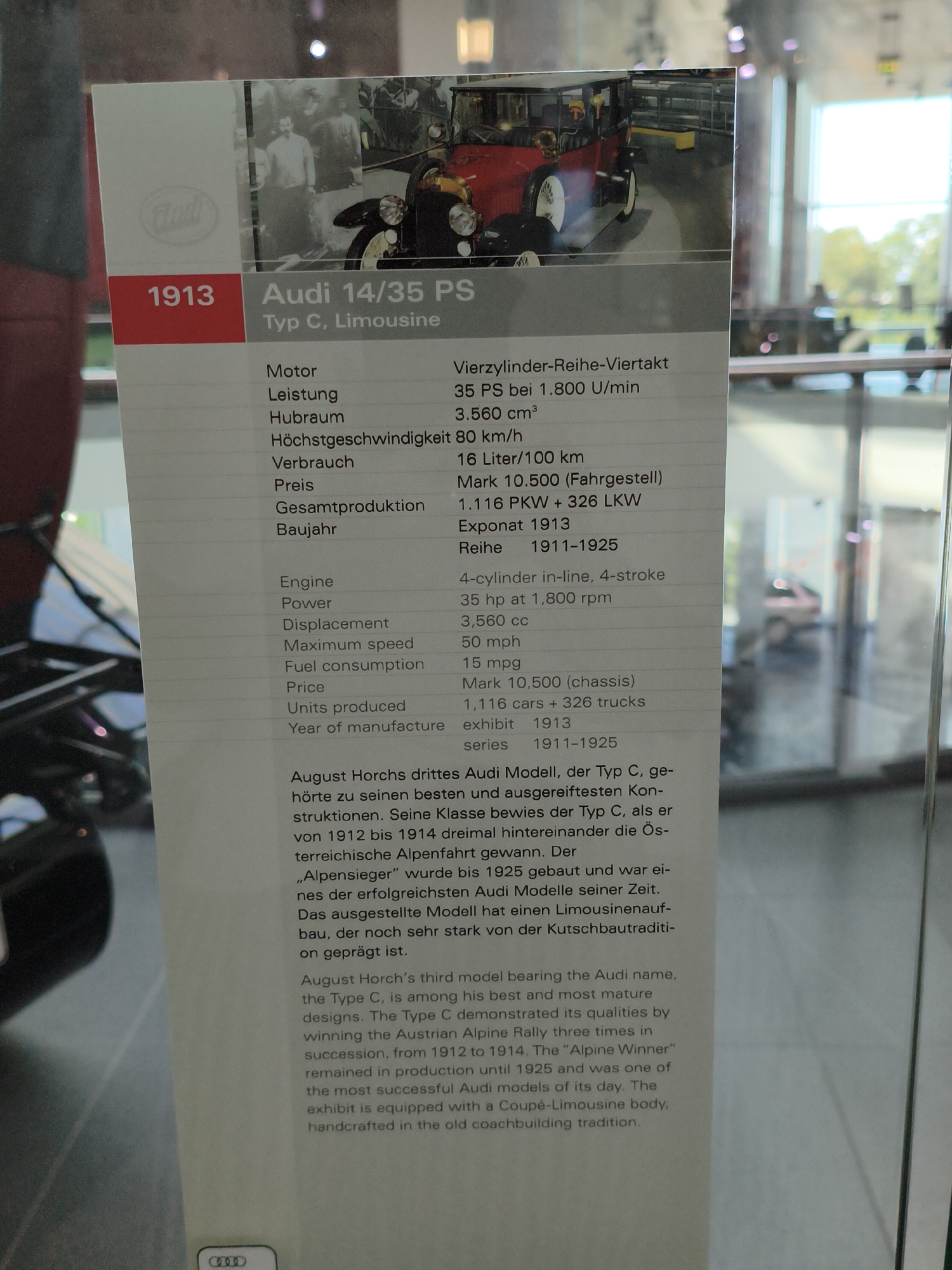 This image displays a placard detailing the Audi 14/35 PS Typ C, Limousine, manufactured in 1913. The placard is situated within a museum setting, likely at the Audi Forum in Ingolstadt, Germany, given the prominent Audi branding and historical context.
The placard features a black and white photograph of the vintage car. To the left, the year "1913" is presented in a red box, adjacent to a faded Audi logo. Below the car image and year, a detailed specification list is provided in two columns. The left column lists categories such as "Motor," "Leistung," "Hubraum," "Höchstgeschwindigkeit," "Verbrauch," "Preis," "Gesamtproduktion," and "Baujahr." The right column provides the corresponding technical data in German, such as "Vierzylinder-Reihe-Viertakt," "35 PS bei 1.800 U/min," "3.560 cm³," "80 km/h," "16 Liter/100 km," "Mark 10.500 (Fahrgestell)," "1.116 PKW + 326 LKW," and "Exponat 1913."
Below this, a similar list is presented in English, with categories like "Engine," "Power," "Displacement," "Maximum speed," "Fuel consumption," "Price," "Units produced," and "Year of manufacture." The English specifications include "4-cylinder in-line, 4-stroke," "35 hp at 1,800 rpm," "3,560 cc," "50 mph," "15 mpg," "Mark 10,500 (chassis)," "1,116 cars + 326 trucks," "exhibit 1913," and "series 1911-1925."
Further down the placard is a narrative description in both German and English. The German text describes August Horch's third Audi model, the Type C, as a successful and mature design, highlighting its performance in the Austrian Alpine Rally three times and its production from 1912 to 1925. The English text reiterates these points, stating that the Type C is among Horch's best and most mature designs, and mentioning its success in the Alpine Rally and its production period. The text also notes that the displayed exhibit is equipped with a coupé-limousine body and is handcrafted in the old coachbuilding tradition.
The background appears to be a museum interior with glass display cases and some reflections of the surrounding environment, including trees and possibly other exhibits. The overall lighting is moderate, with some light sources visible as blurred orbs in the upper part of the image. No people are clearly visible in the foreground or interacting with the placard or car.