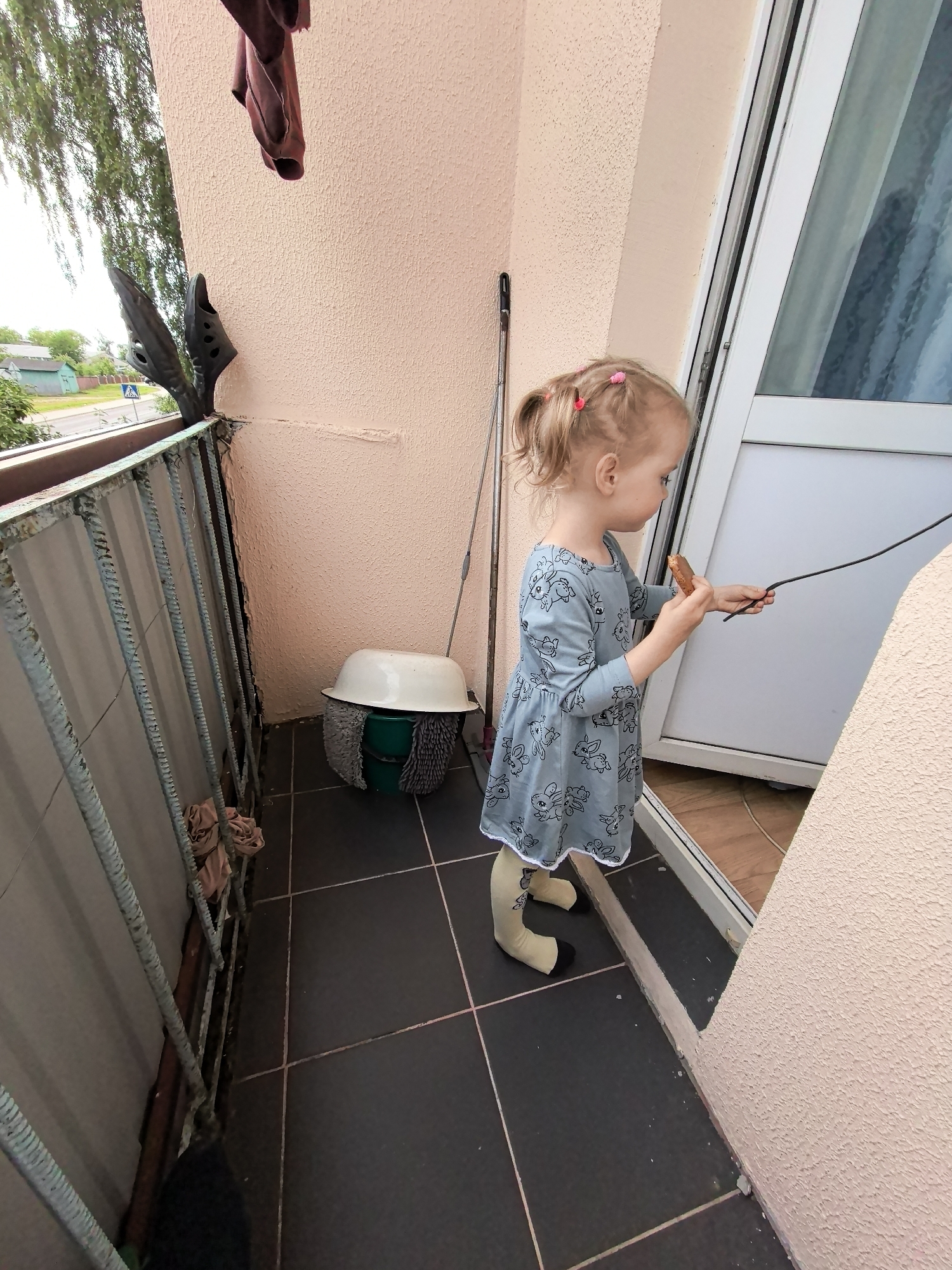 A blonde child with two blonde ponytails, wearing a light blue dress with a bunny pattern, stands on a balcony. The child is holding a cookie and a thin black rod. The balcony floor is tiled in dark grey with pink grout lines. To the left of the child, a metal railing is visible with some brown fabric draped over it. Behind the child, a white basin sits atop a green bucket, with a grey mop head around the bucket. A mop is leaning against the wall behind the basin. The wall of the balcony is a textured pale pink. A white doorframe with a glass panel is to the child's right. In the background, trees and houses are visible, suggesting an urban or suburban setting. There are also a pair of black sandals placed on top of the railing on the far left.