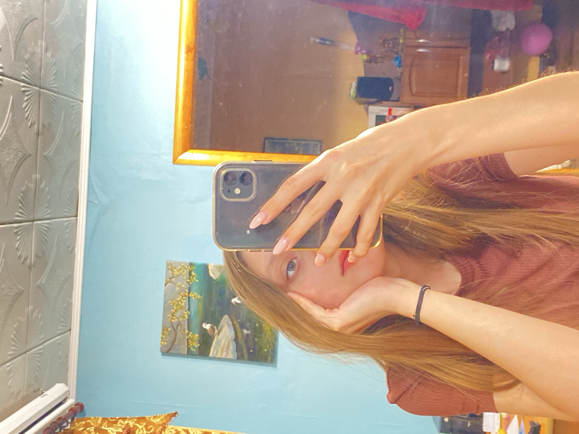 A selfie shows a person with long blonde hair and fair skin, wearing a maroon top and a black band on their left wrist. They are holding a black iPhone in front of their face with one hand, their cheek resting on the other hand. The background includes a bathroom with light blue walls, ornate grey tiles on the left, and a framed painting of trees on the wall. A mirror with a wooden frame is also visible, reflecting a bedroom or living area with furniture and decorations. The location is Unknown, Unknown.